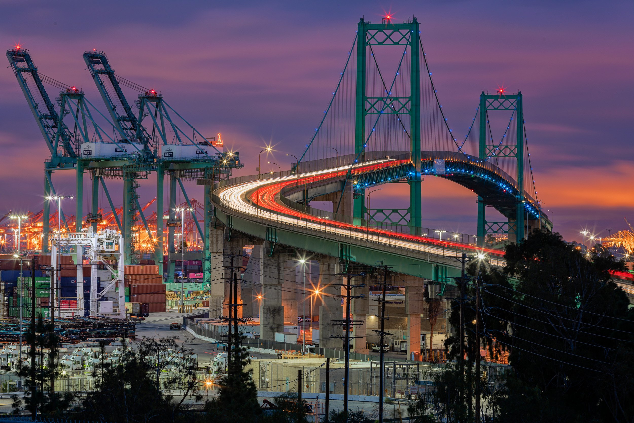 Horizontal long-exposure photo of traffic light trails winding across the illuminated Vincent Thomas Bridge at twilight with the Port of Los Angeles cranes in the background.