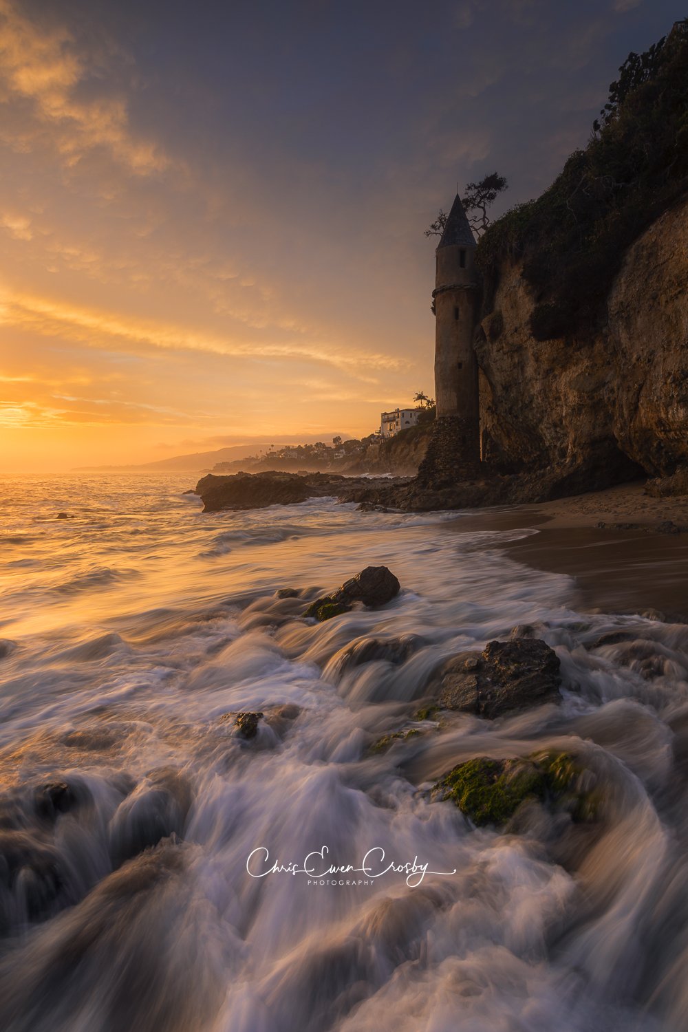 A vertical photograph of the stone Victoria Beach Pirate Tower in Laguna Beach, California, during a vibrant sunset with pink and orange clouds and moving tide at the base.