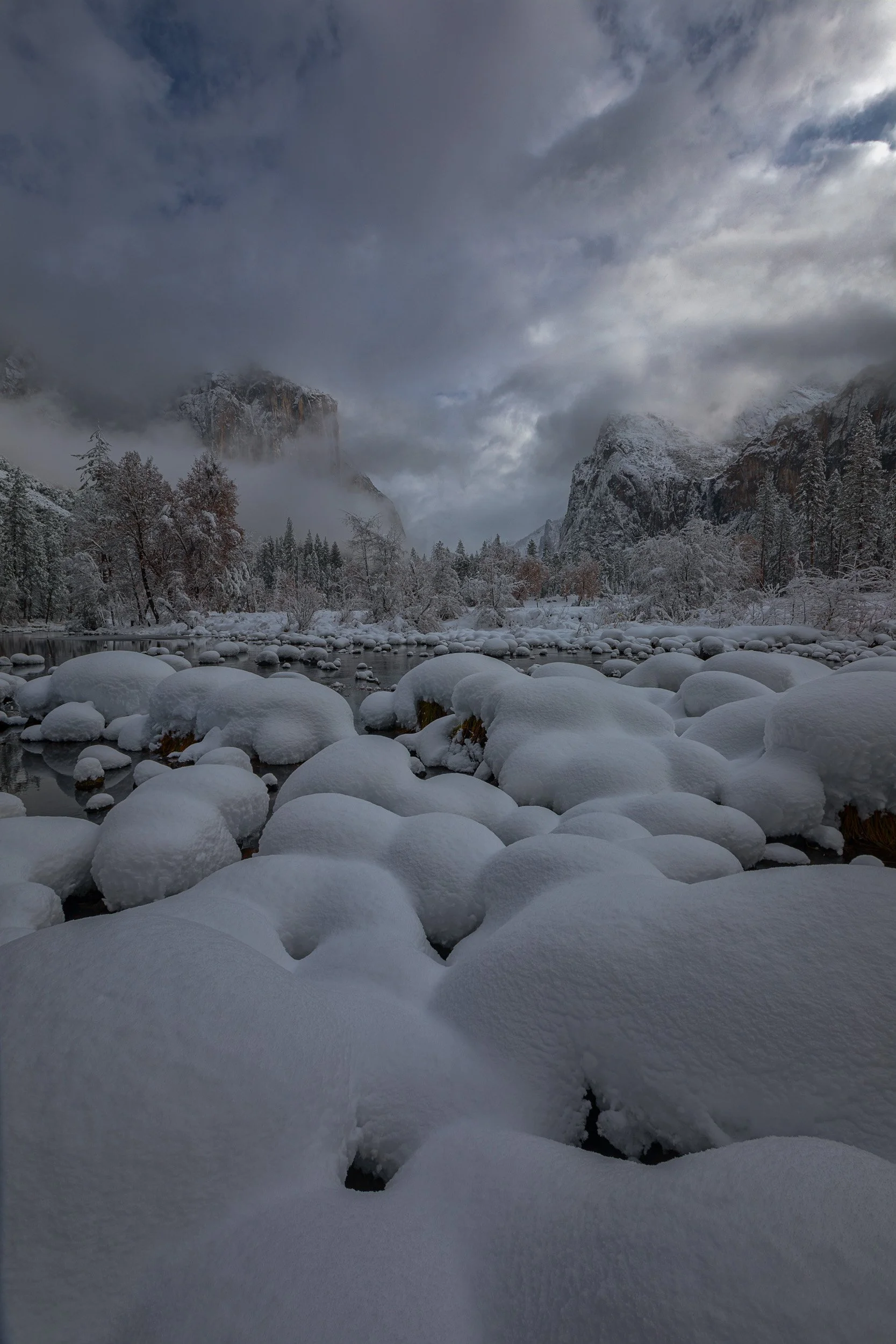 Vertical winter landscape photograph of soft mounds of fresh snow along the Merced River looking toward fog-shrouded granite cliffs at Valley View in Yosemite National Park by Chris Ewen Crosby.