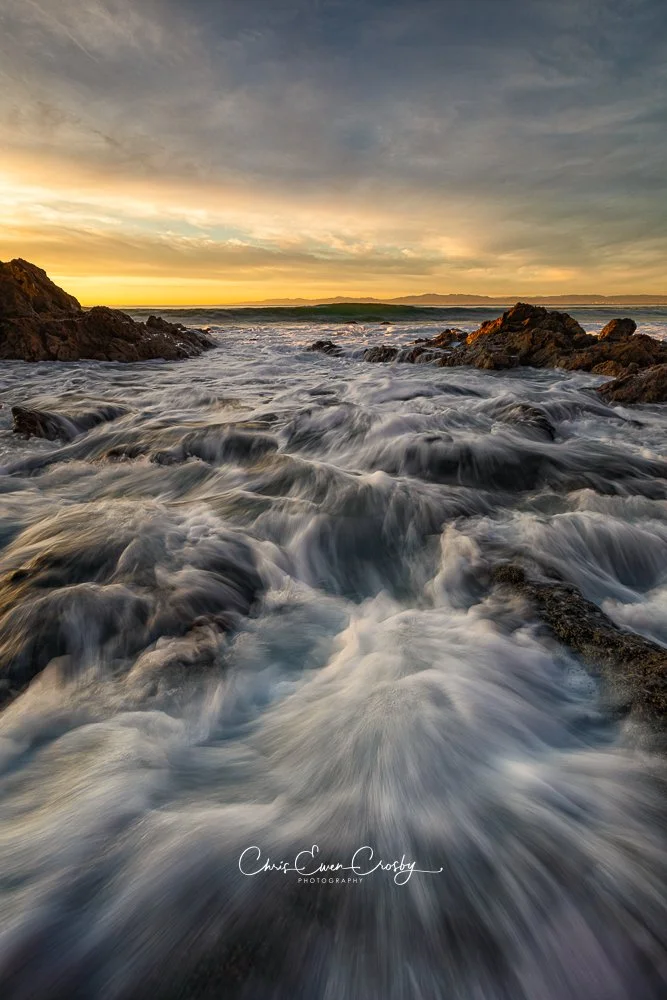 Vertical landscape photo of waves rushing over dark rocks at Palos Verdes during golden hour, creating white frothy water textures.