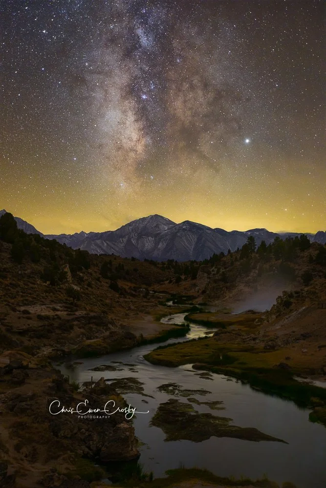 A vertical night photograph of the Milky Way galaxy stretching over a winding creek with steam rising from geothermal vents at Hot Creek, California.