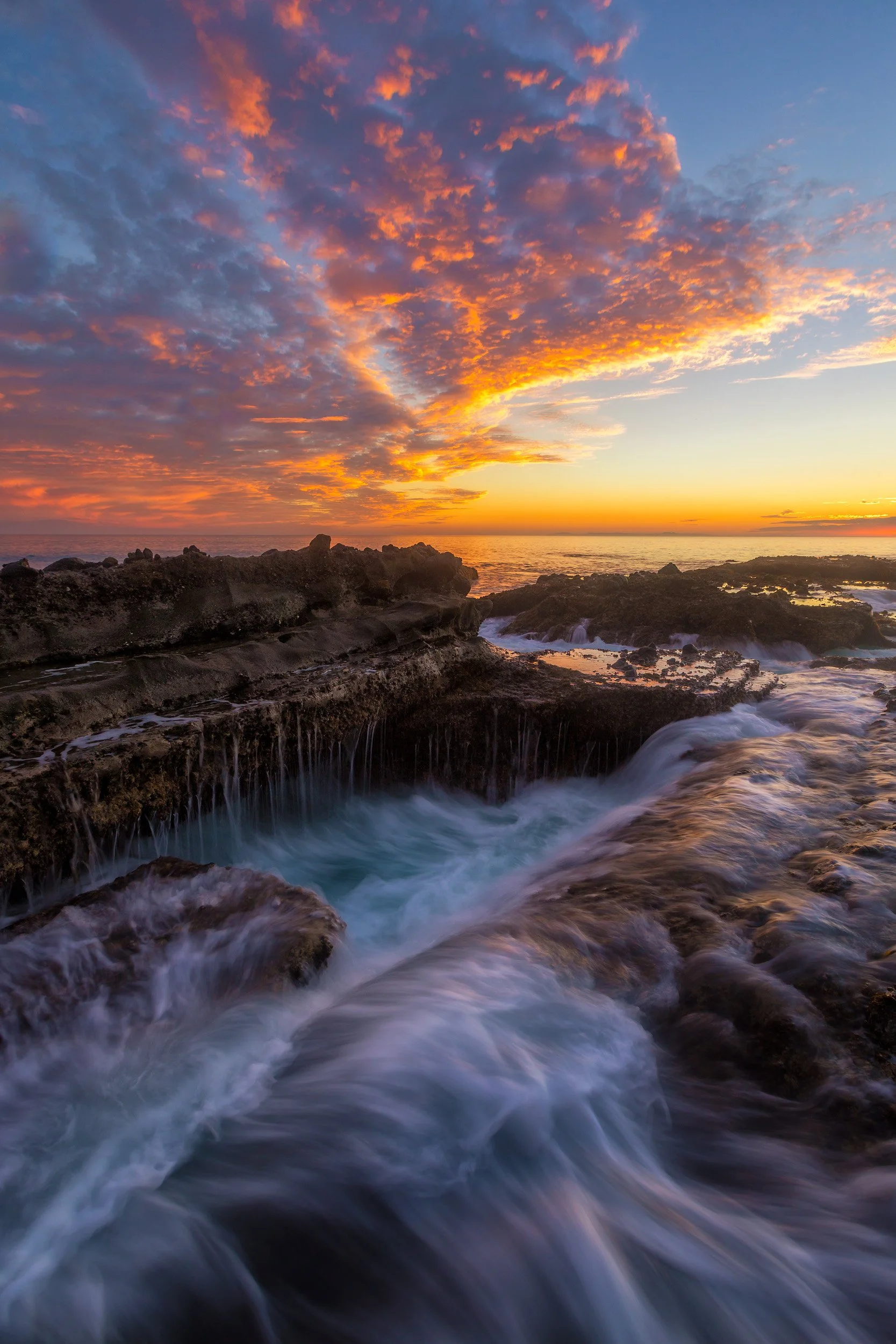 Vertical long exposure of waves cascading over rocky tide pool ledges under a dramatic orange sunset sky in Laguna Beach by Chris Ewen Crosby.