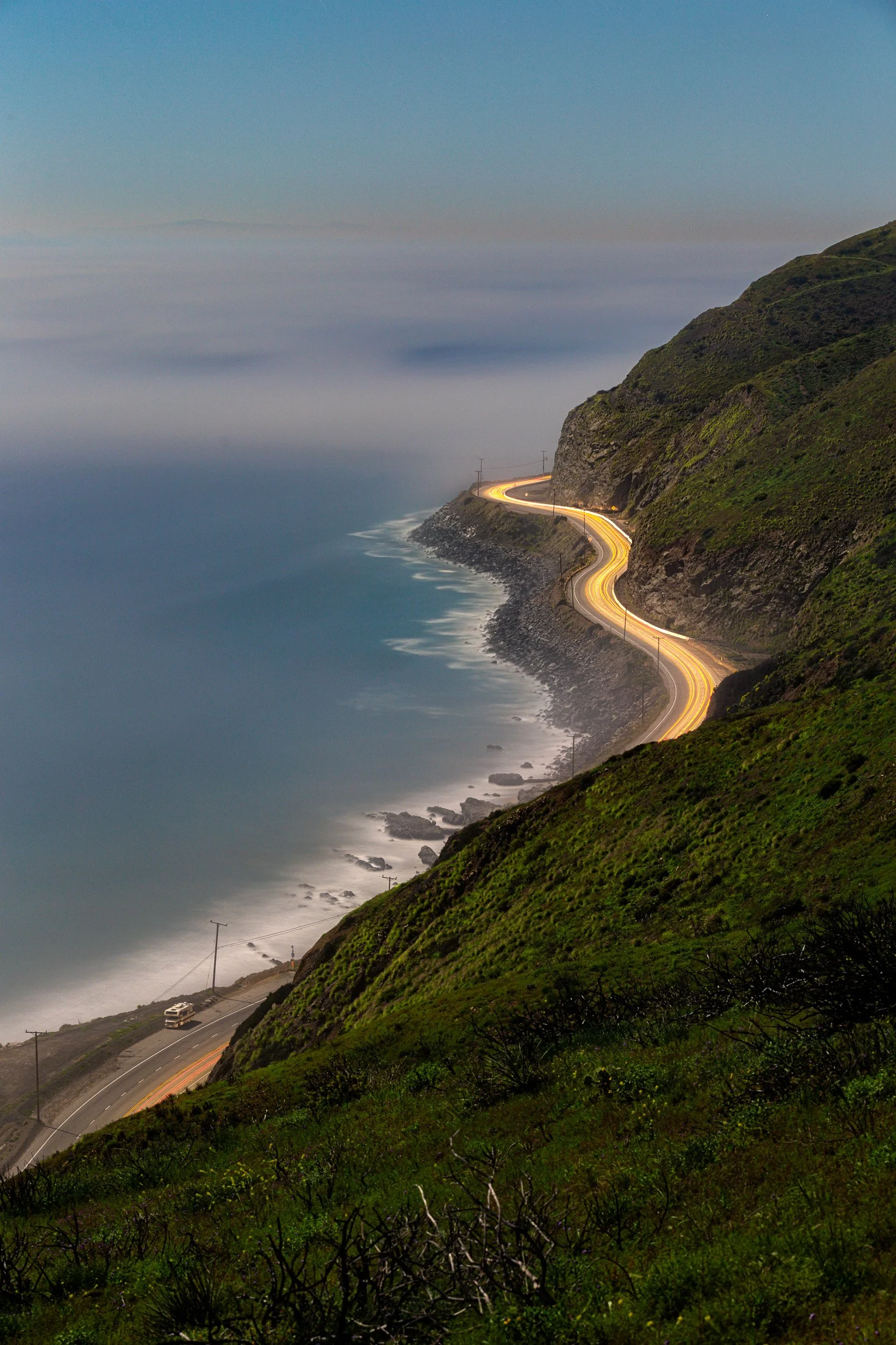 Vertical night photography photograph of glowing headlight light trails winding along Pacific Coast Highway beside rocky ocean cliffs in Southern California by Chris Ewen Crosby.