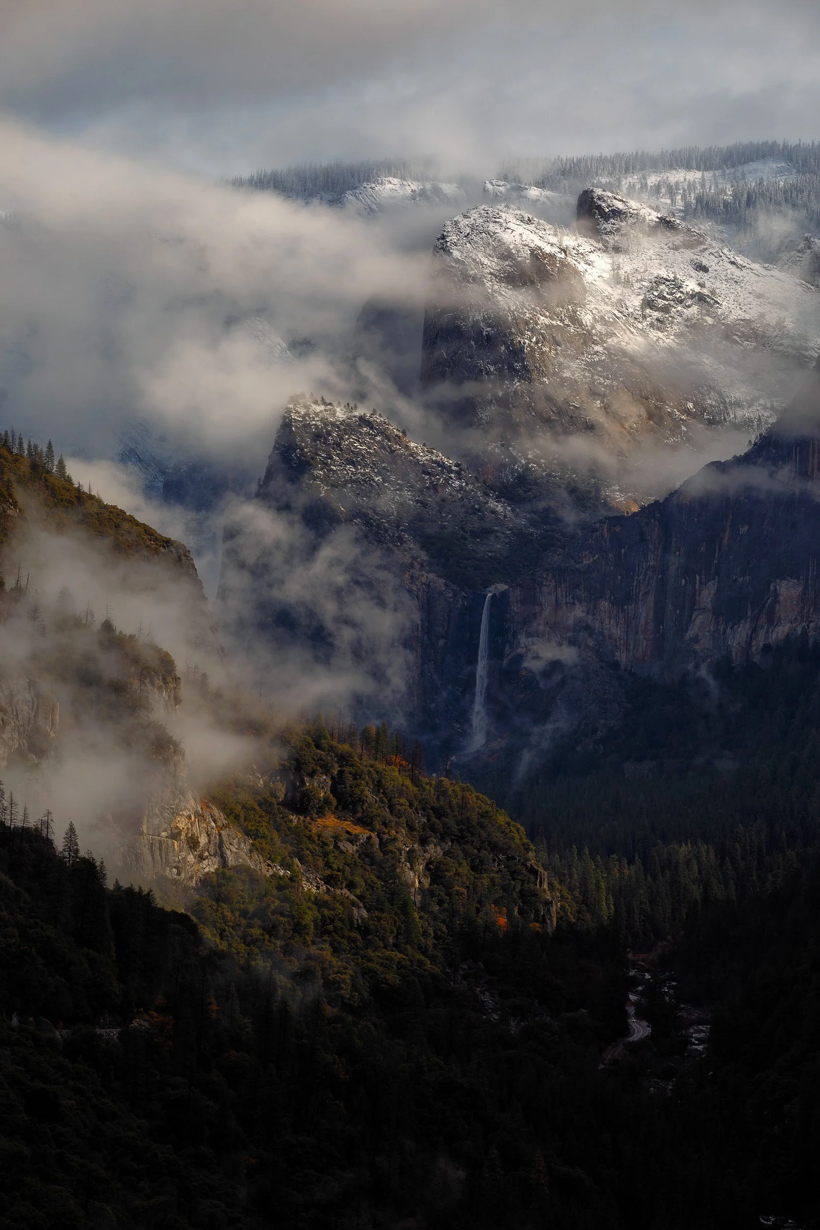 Swirling valley fog surrounds a distant waterfall and snow-dusted granite peaks under shifting sunlight in Yosemite.
