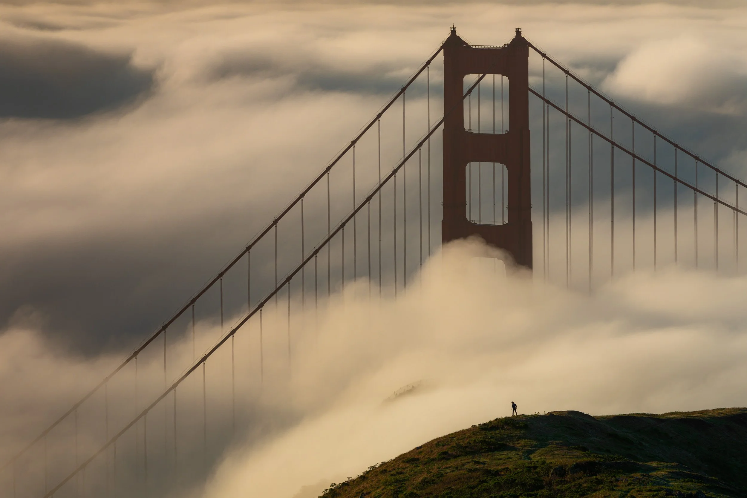 Silhouette of a person standing on a grassy hill overlooking the Golden Gate Bridge tower enveloped in thick fog by Chris Ewen Crosby.