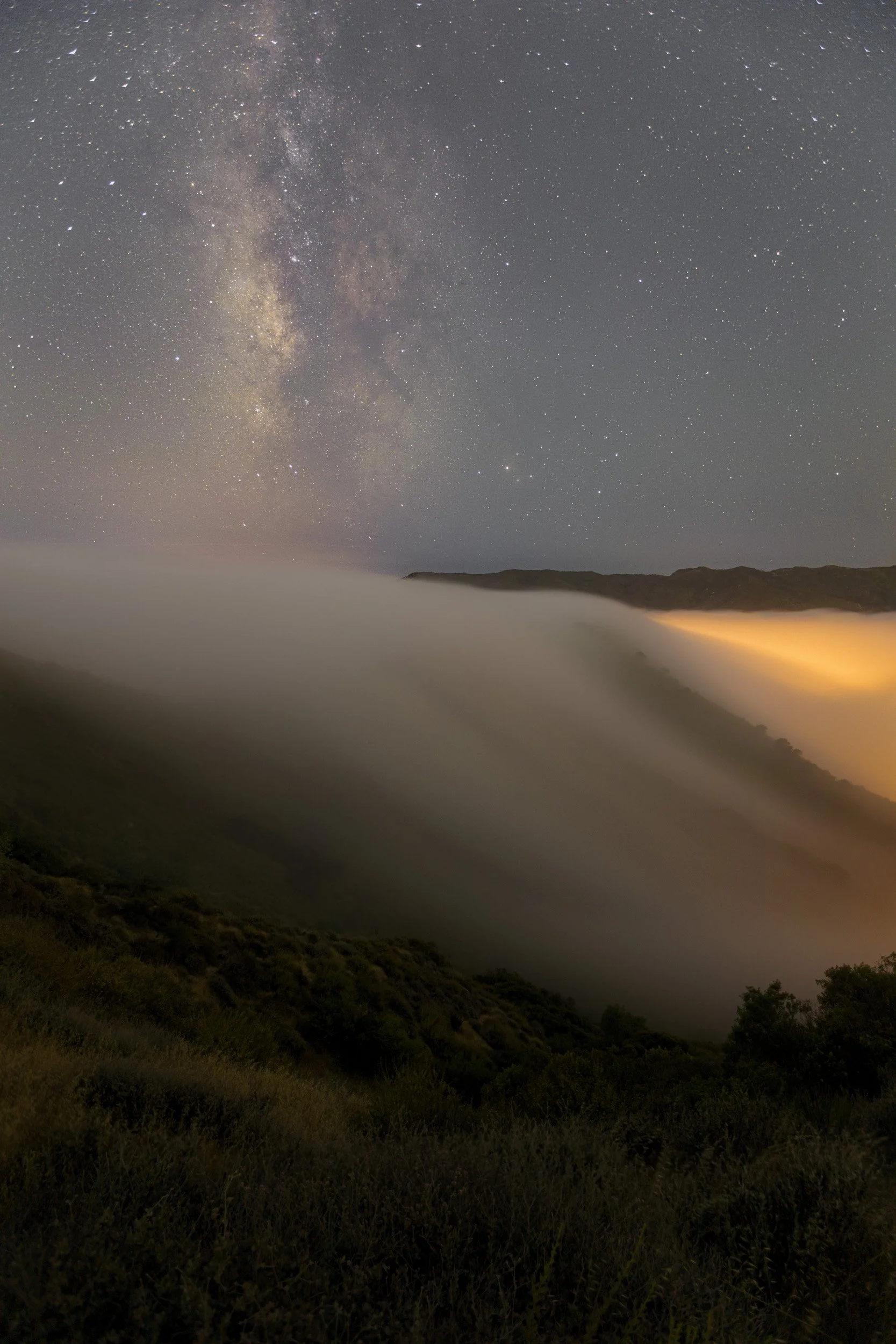 Vertical night astrophotography photograph of dense coastal marine layer fog rolling over mountain ridges under the Milky Way galaxy in the Santa Monica Mountains by Chris Ewen Crosby.