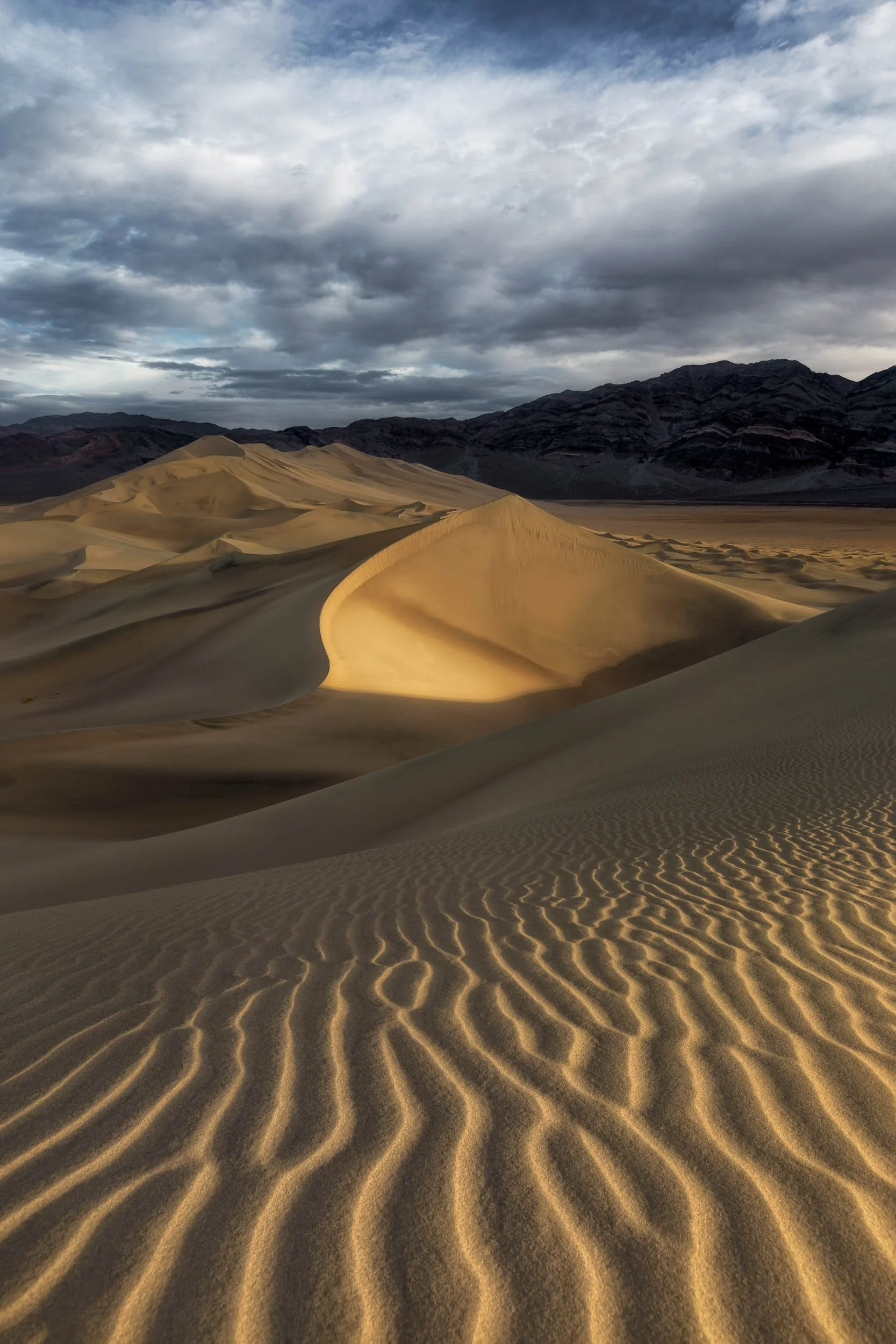 Close-up of wind-blown sand ripples leading to a large sunlit dune with rugged mountains under a cloudy sky in Death Valley.