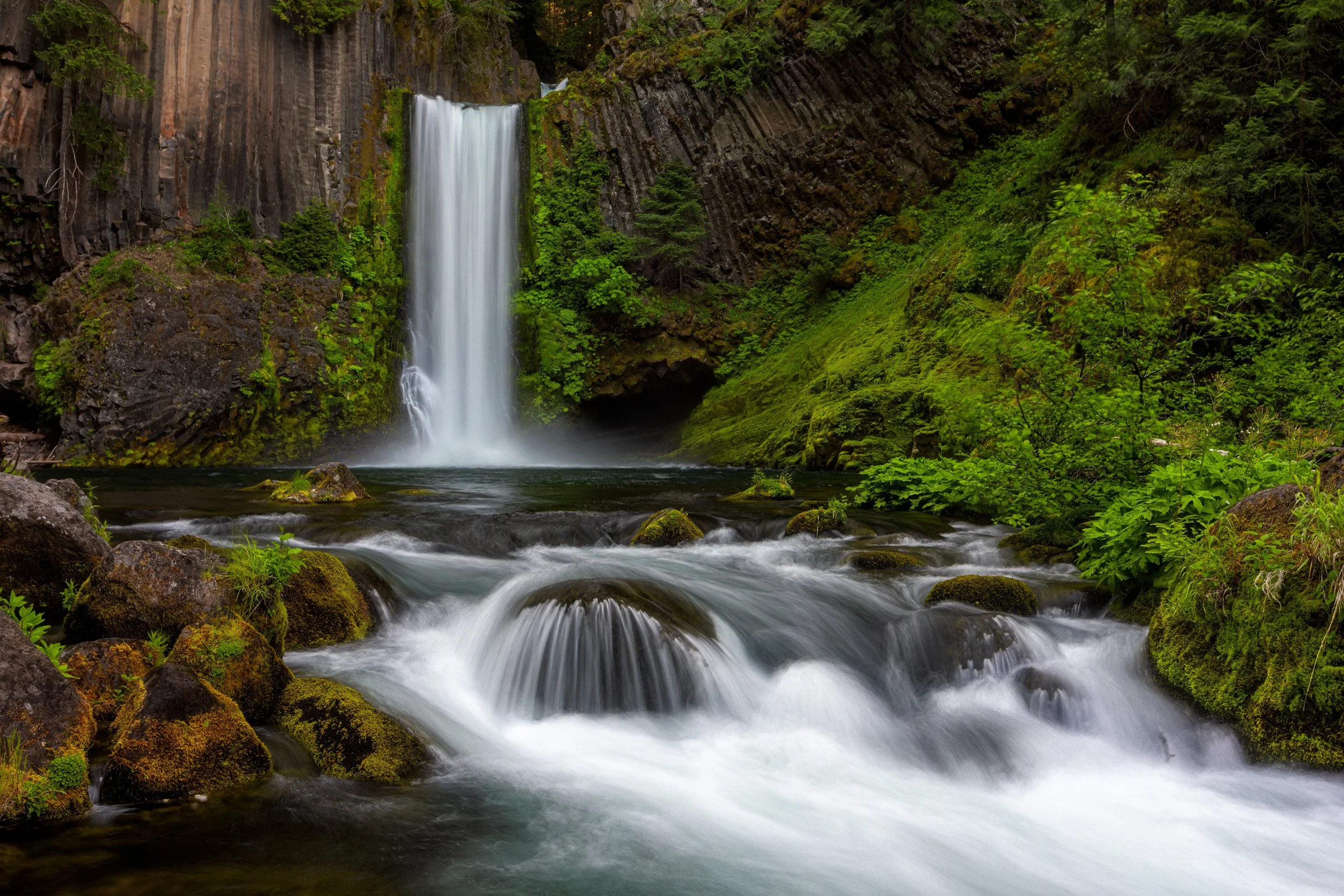 Horizontal long exposure photograph of Toketee Falls plunging over ancient columnar basalt cliffs into a lush, mossy river gorge in Oregon by Chris Ewen Crosby.