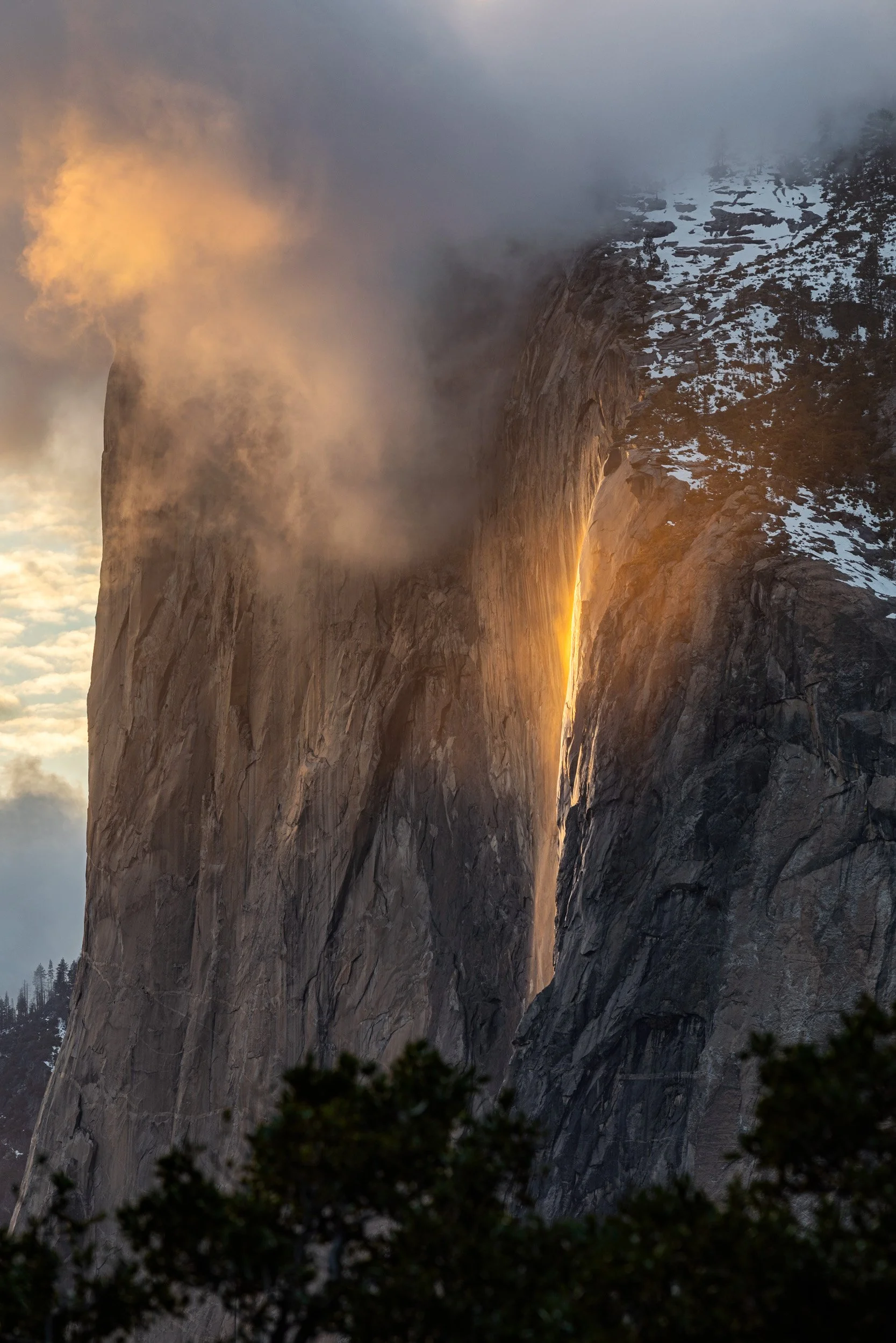Vertical landscape photograph of Horsetail Fall glowing vibrant orange like lava down the granite wall of El Capitan with dramatic sunset clouds in Yosemite National Park by Chris Ewen Crosby.