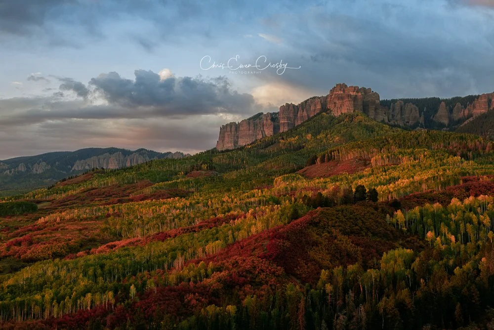 Wide panoramic sunset view of Cathedral Rock in Colorado with red fall foliage, green aspens, and a snow-dusted mountain peak.