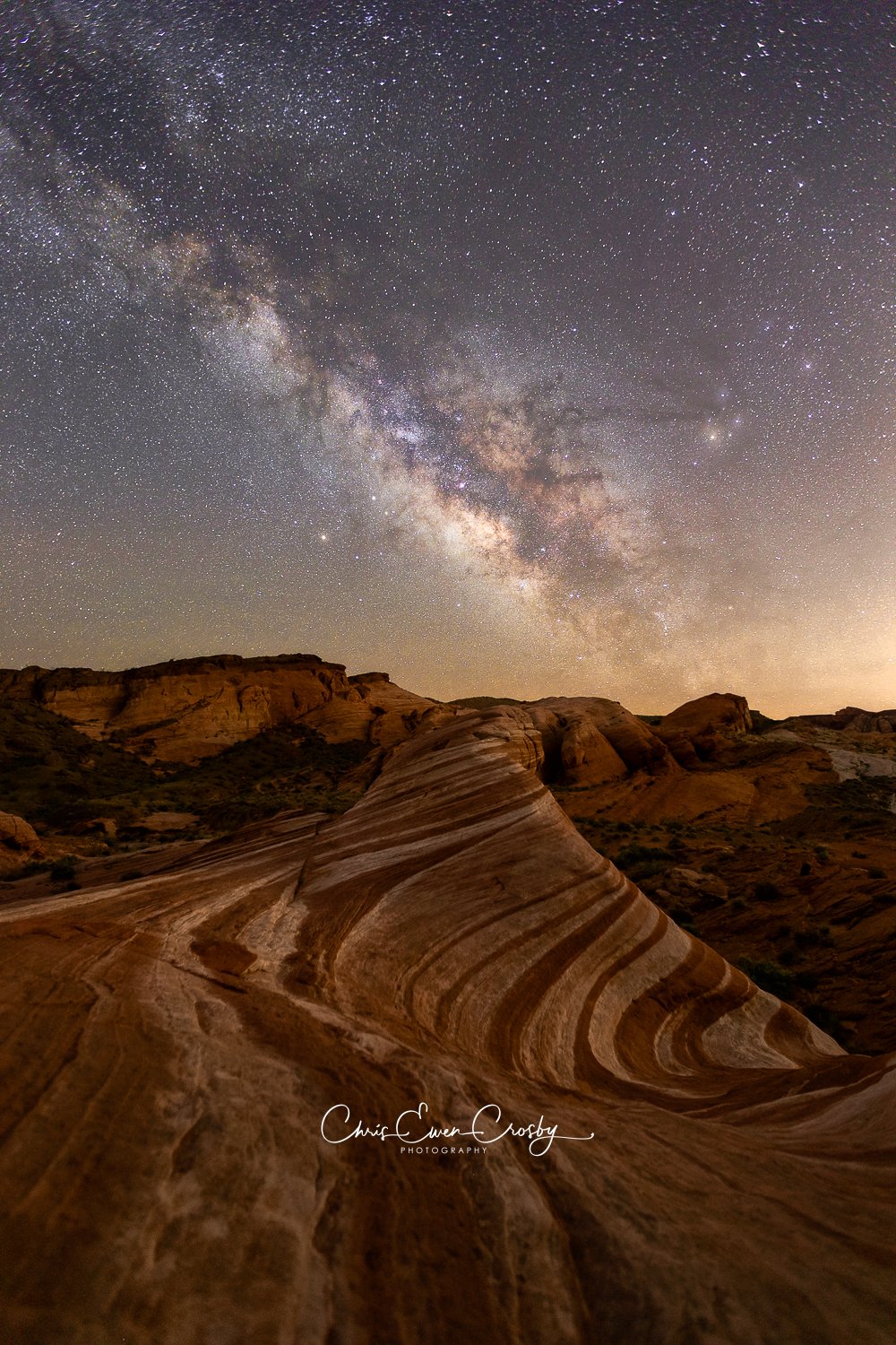 A vertical night photograph titled "Chocolate Milky Way" showing the Milky Way galaxy in a starry sky above swirling red and white sandstone rock formations in Valley of Fire State Park.