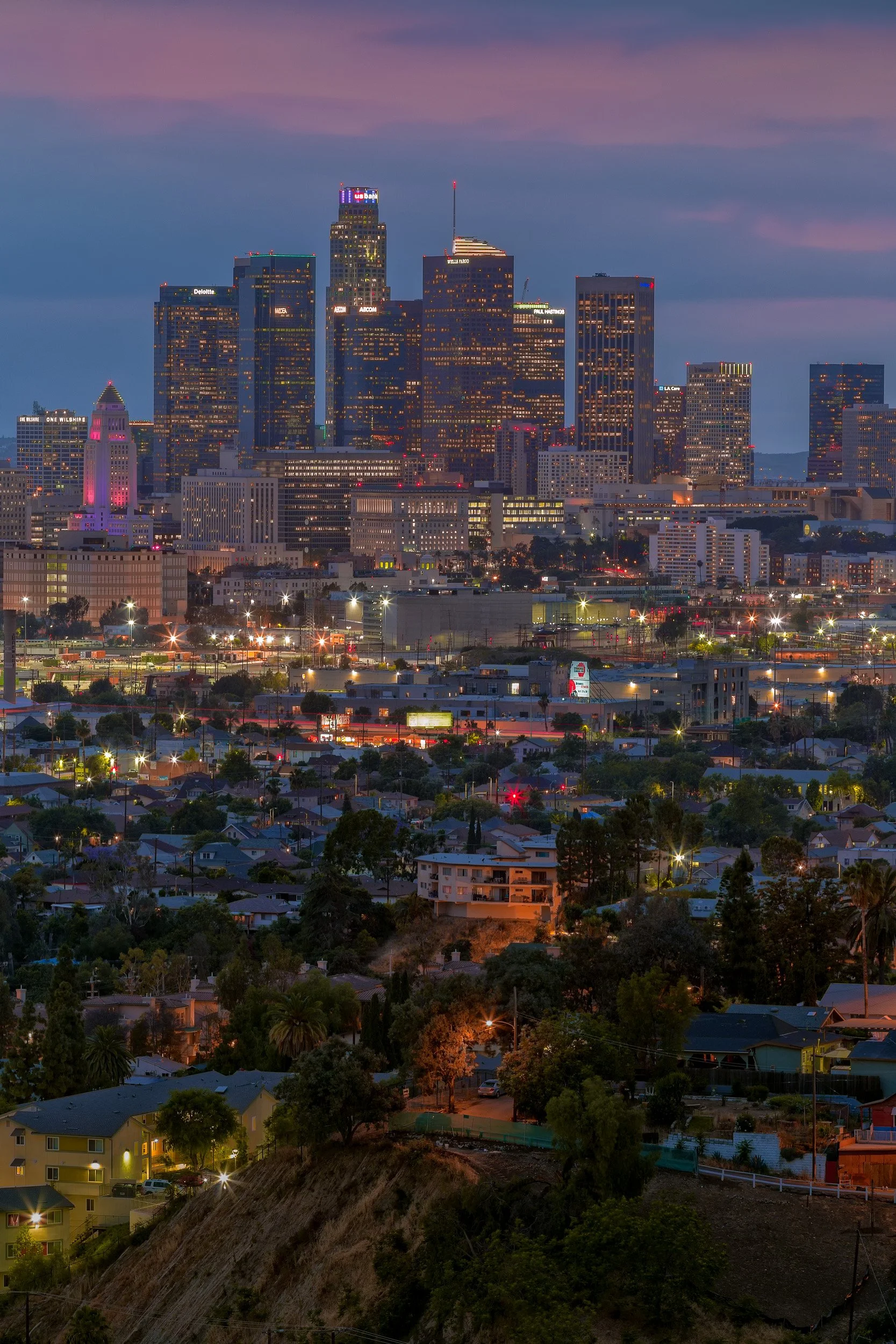 Vertical twilight cityscape of the Downtown Los Angeles skyline with glowing skyscrapers and city lights under a purple dusk sky.