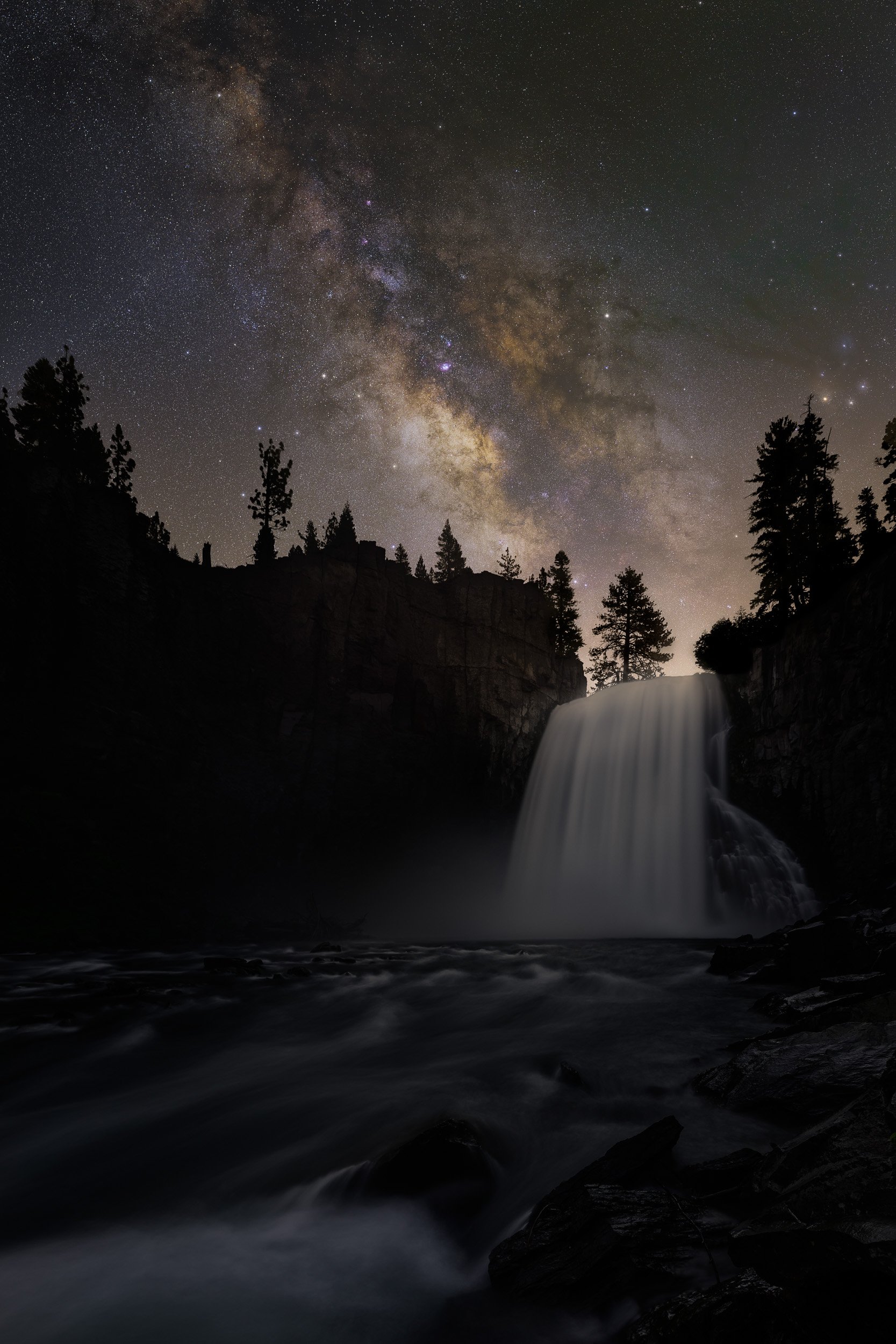 Vertical night astrophotography photograph of a rushing waterfall silhouetted against pine trees and the bright Milky Way galaxy rising overhead in Mammoth Lakes, California by Chris Ewen Crosby.