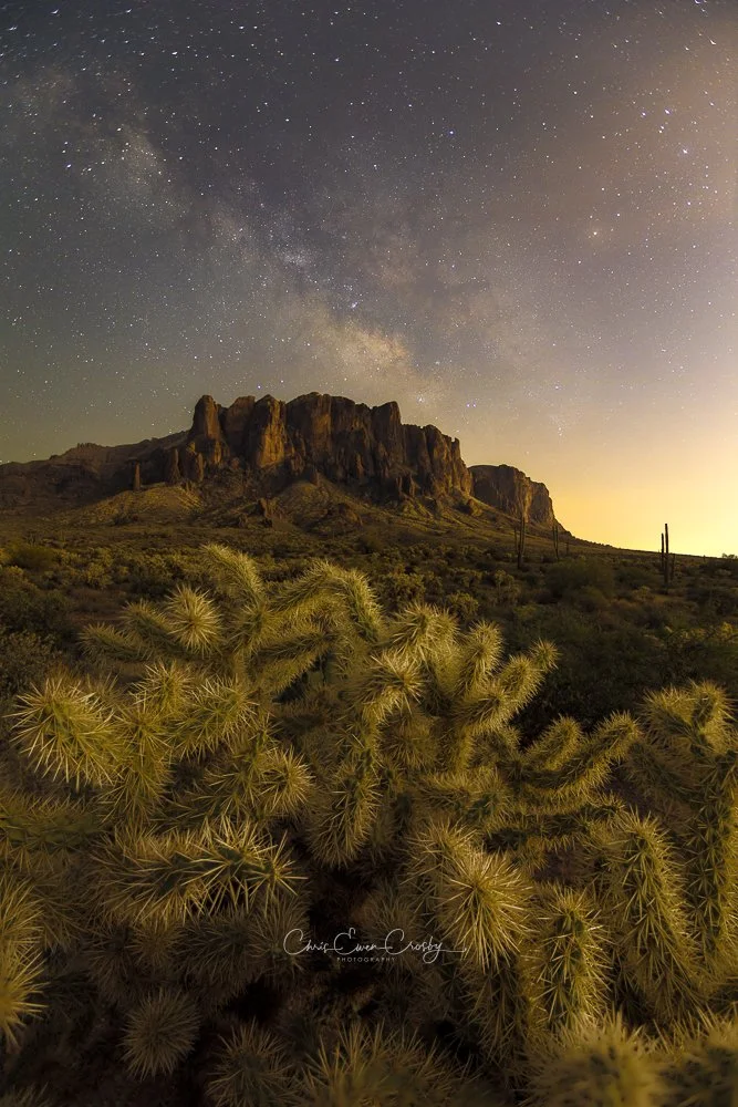 Vertical long-exposure photograph of the Milky Way over the Superstition Mountains with a glowing Cholla cactus in Lost Dutchman State Park, Arizona.