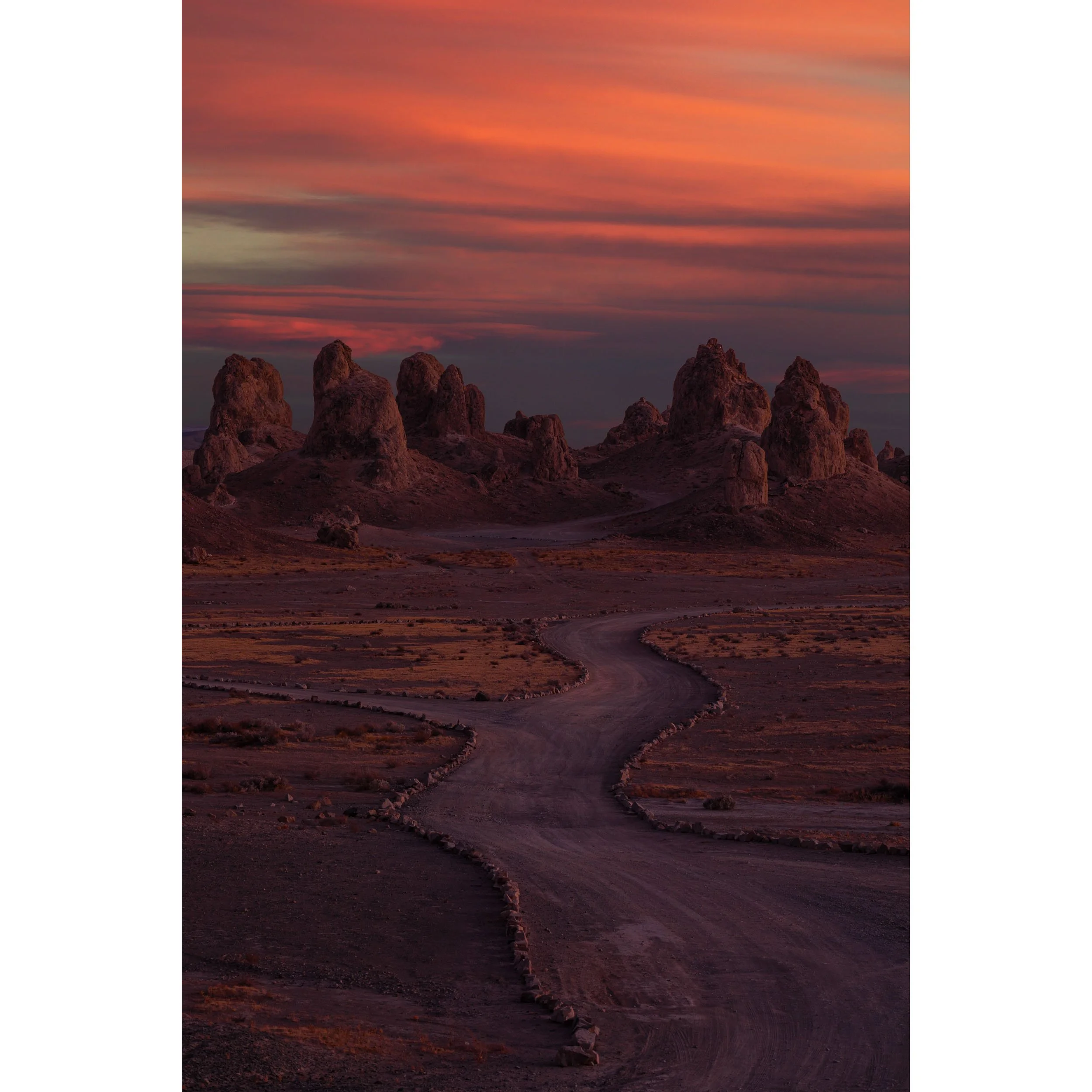 Vertical fine art landscape photograph of a dirt road winding toward the tufa spires of Trona Pinnacles under a fiery red sunset sky.