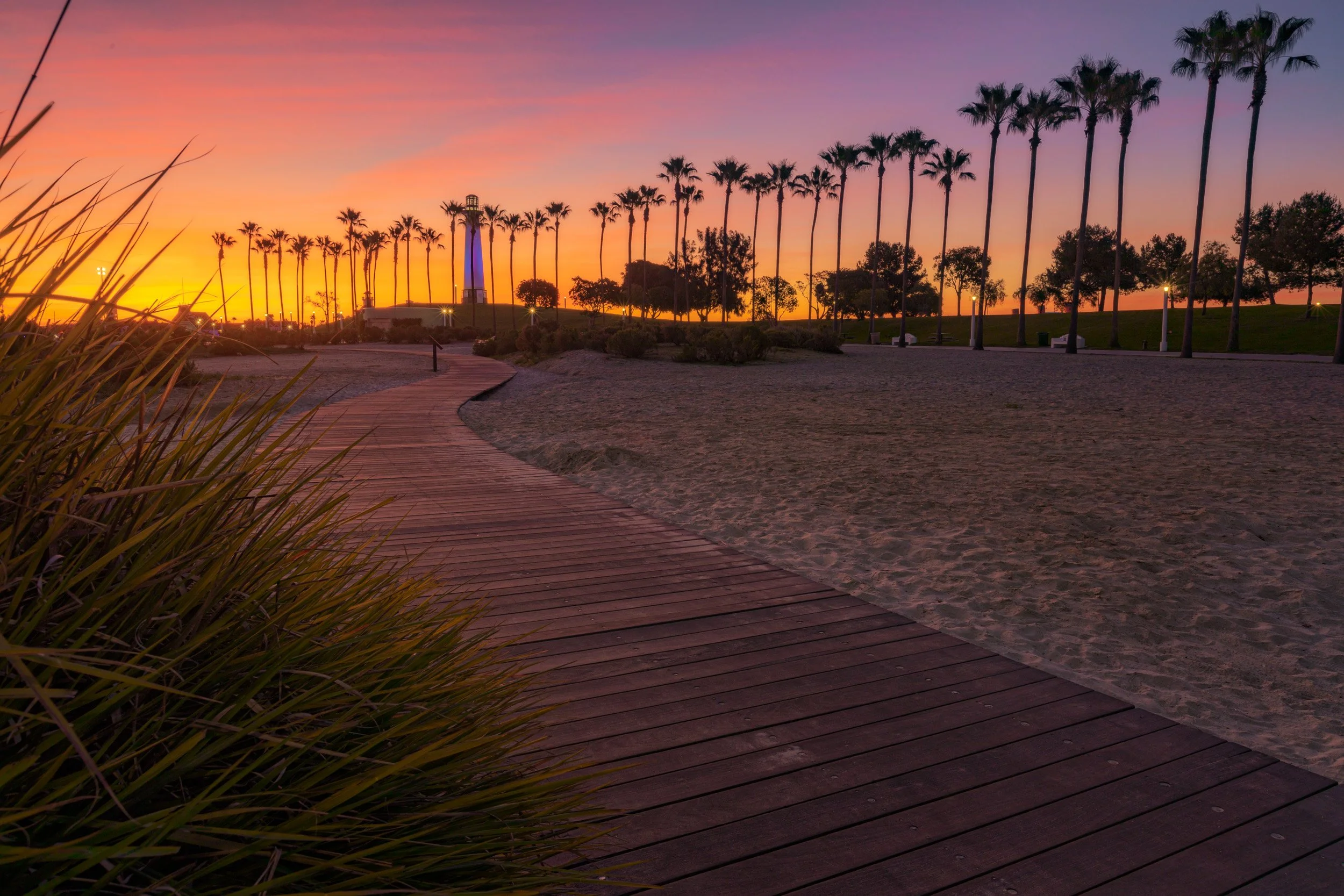 Curving wooden boardwalk leading toward the illuminated Lions Lighthouse at Rainbow Lagoon Park in Long Beach, California at sunrise by Chris Ewen Crosby.