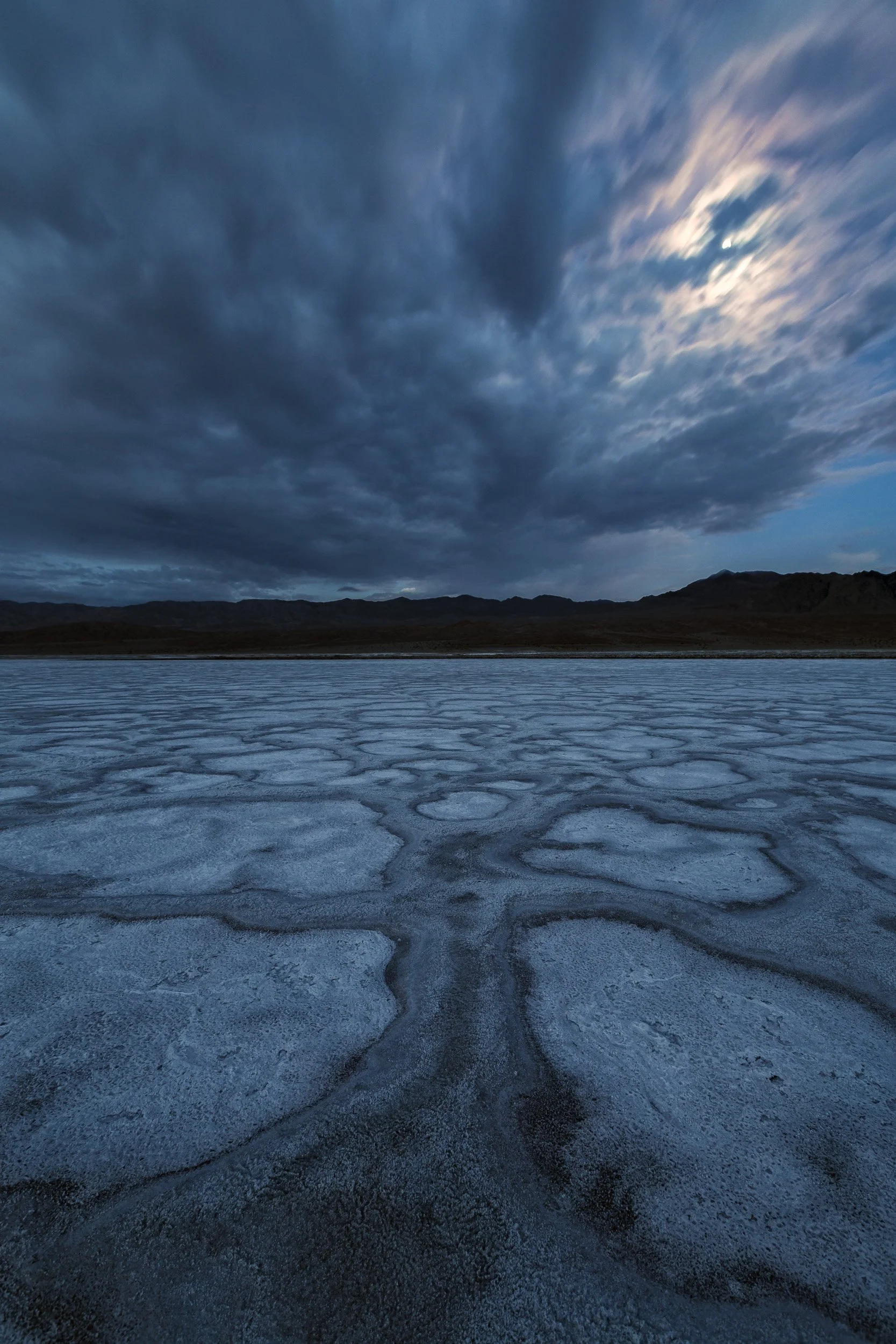 Vertical night photography photograph of expansive salt polygon patterns stretching toward distant mountains under a stormy moonlit sky in Death Valley National Park by Chris Ewen Crosby.