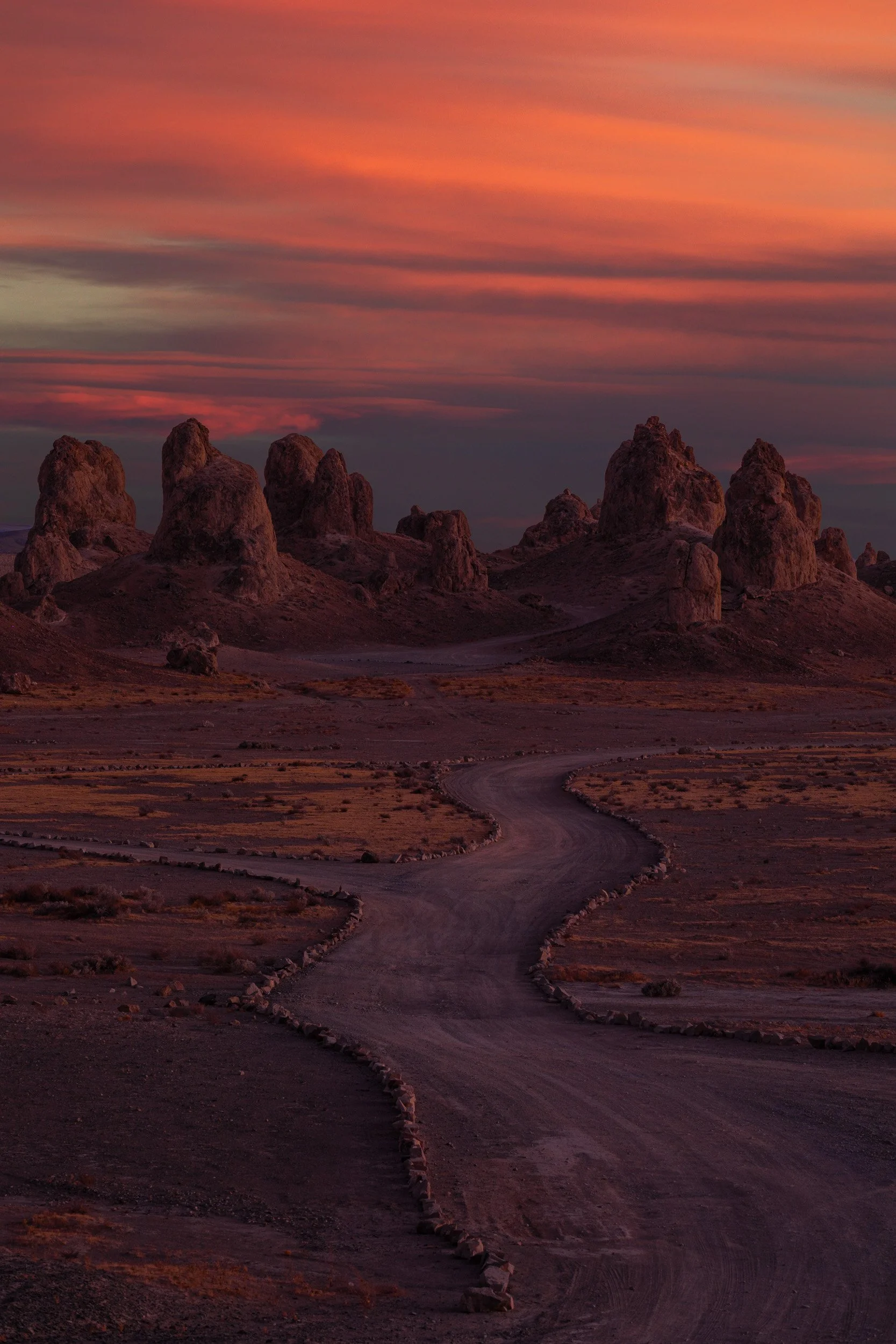 Vertical landscape photograph of a winding dirt road leading toward the tufa spires of Trona Pinnacles under a crimson twilight sunset sky by Chris Ewen Crosby.