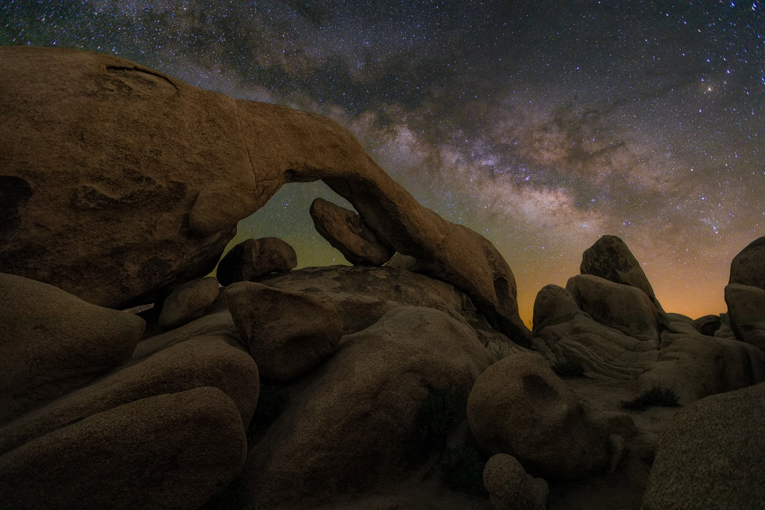 Horizontal night astrophotography photograph of Arch Rock framed against the glowing Milky Way galaxy and starry sky in Joshua Tree National Park by Chris Ewen Crosby.