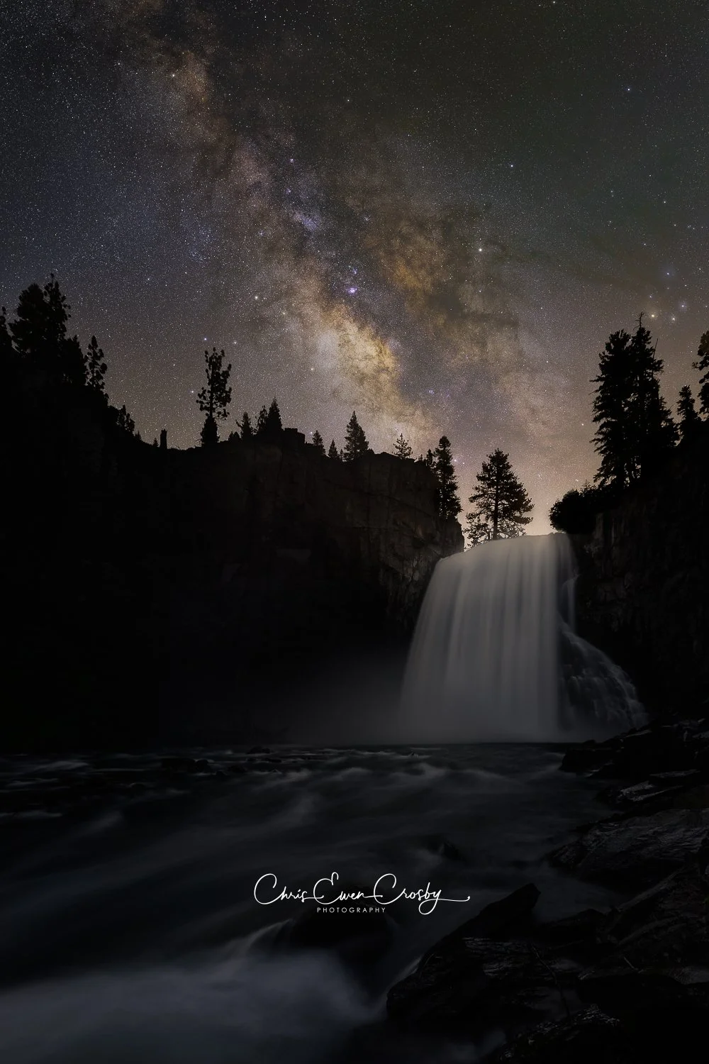 A high-resolution vertical night photograph of Rainbow Falls in Mammoth Lakes with the Milky Way galaxy core glowing intensely above the rushing waterfall and mist.