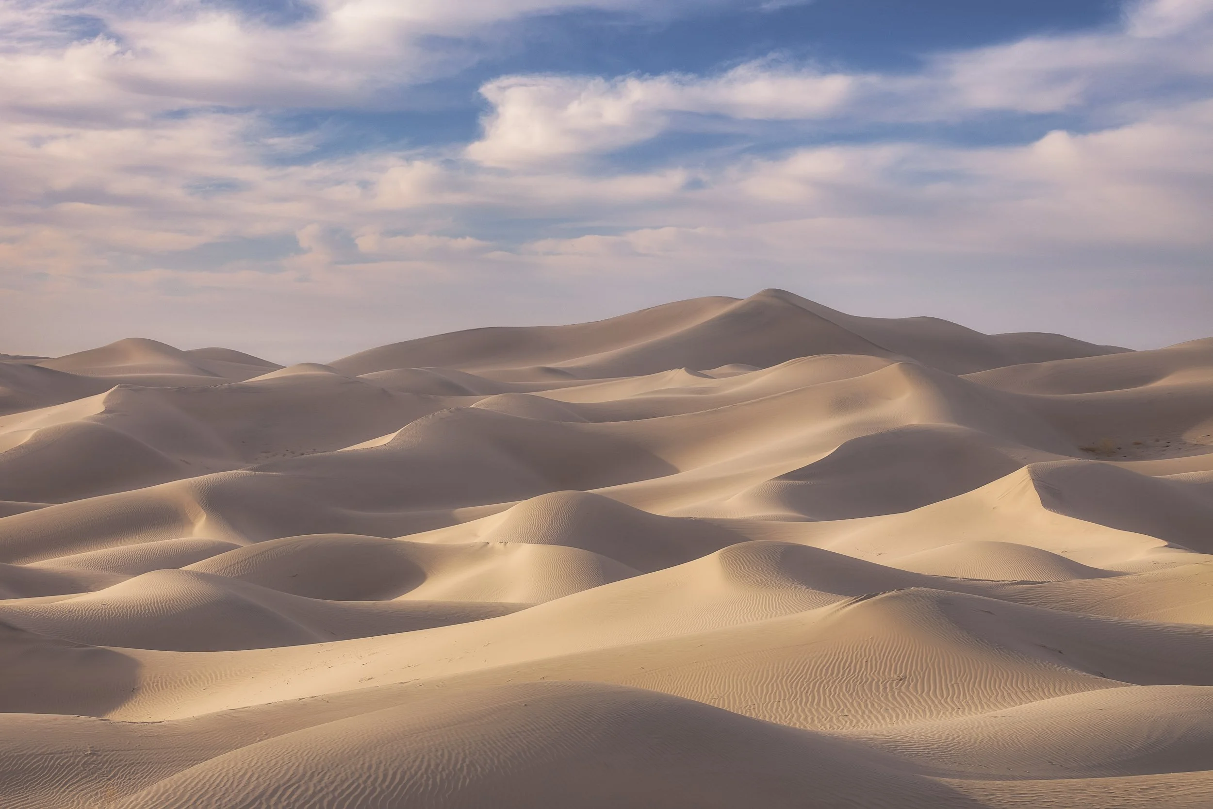 Rolling, golden sand dunes with soft shadows under a bright blue sky with wispy clouds in Death Valley.