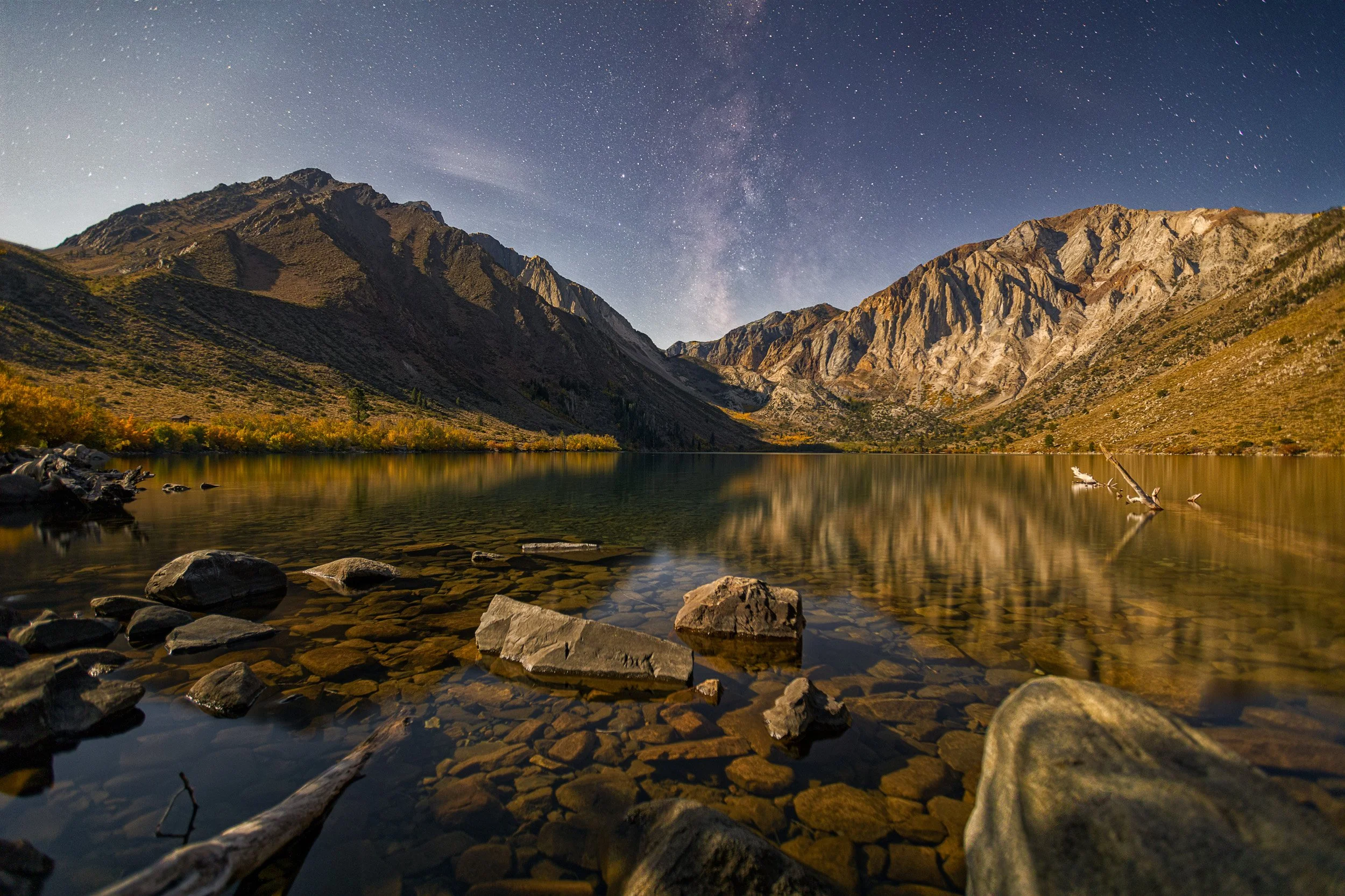 Horizontal fine art night landscape photo of low moonlight illuminating the autumn mountains and lake reflection at Convict Lake under the Milky Way.