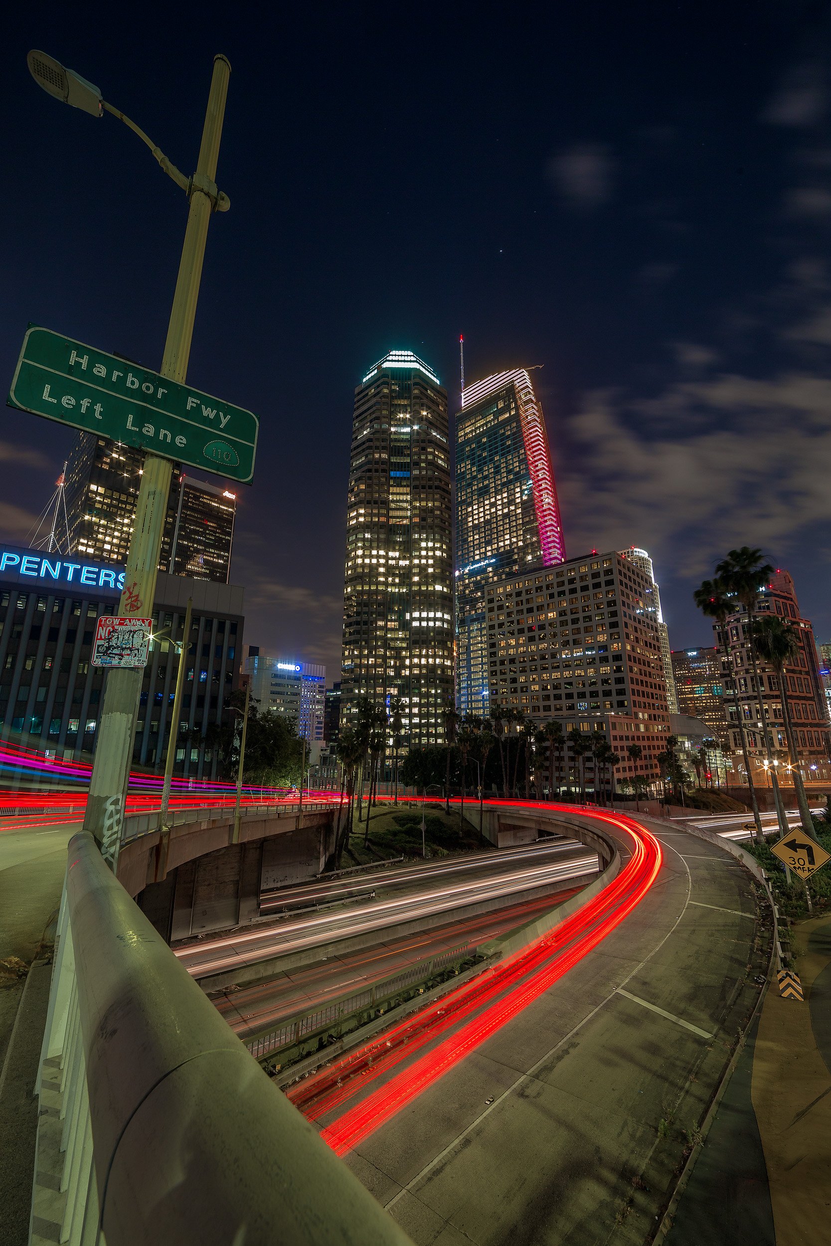 Long exposure light trails curving onto the Harbor Freeway 110 onramp beneath illuminated Downtown Los Angeles skyscrapers by Chris Ewen Crosby.