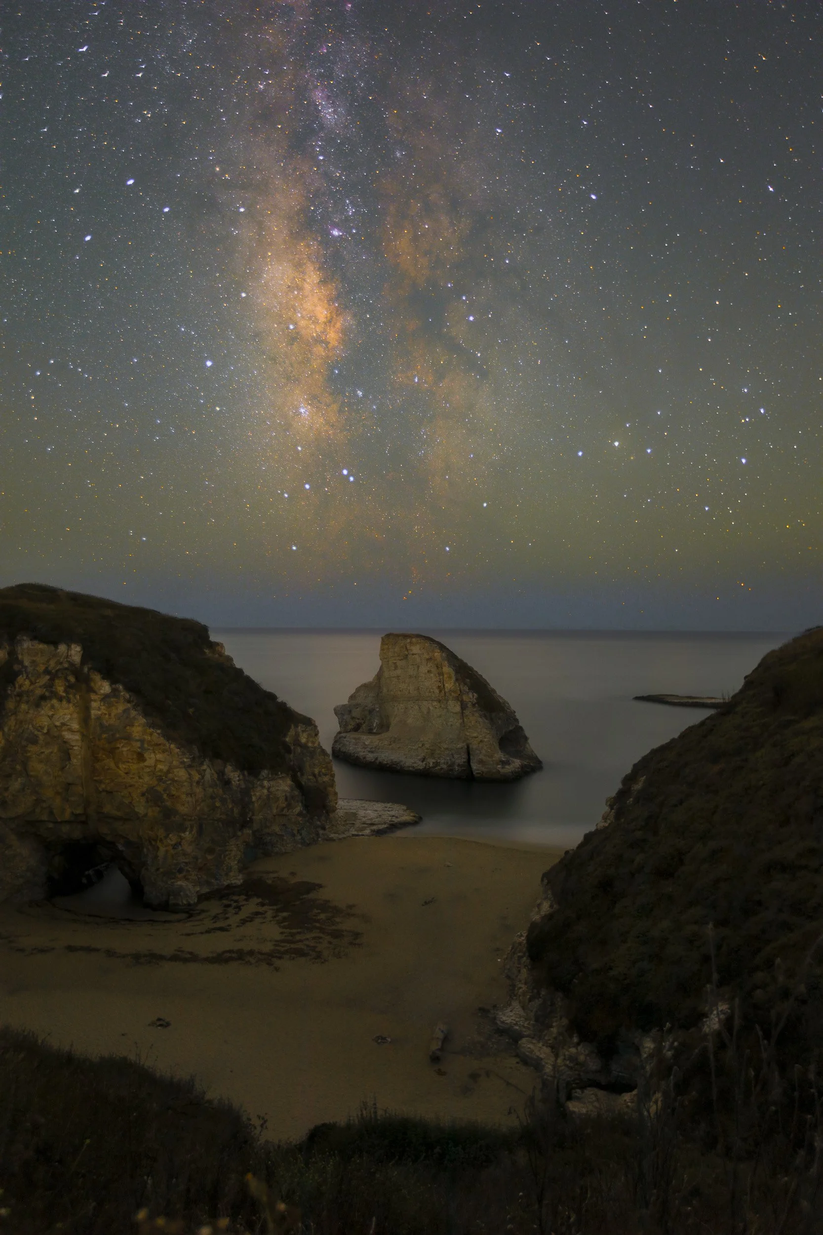 Vertical night astrophotography photograph of the famous Shark Fin sea stack framed by coastal cliffs and the vertical Milky Way galaxy core at Shark Fin Cove near Davenport, California by Chris Ewen Crosby.