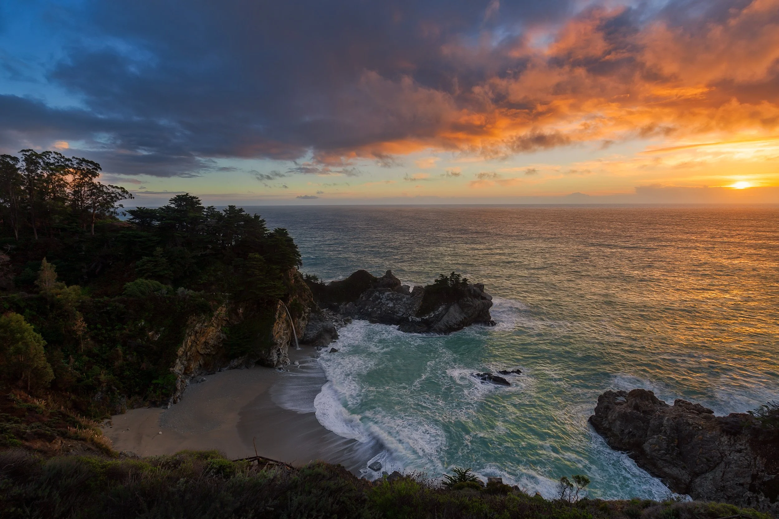 Horizontal landscape photograph of McWay Falls cascading onto a pristine sandy cove during a dramatic sunset in Big Sur by Chris Ewen Crosby.