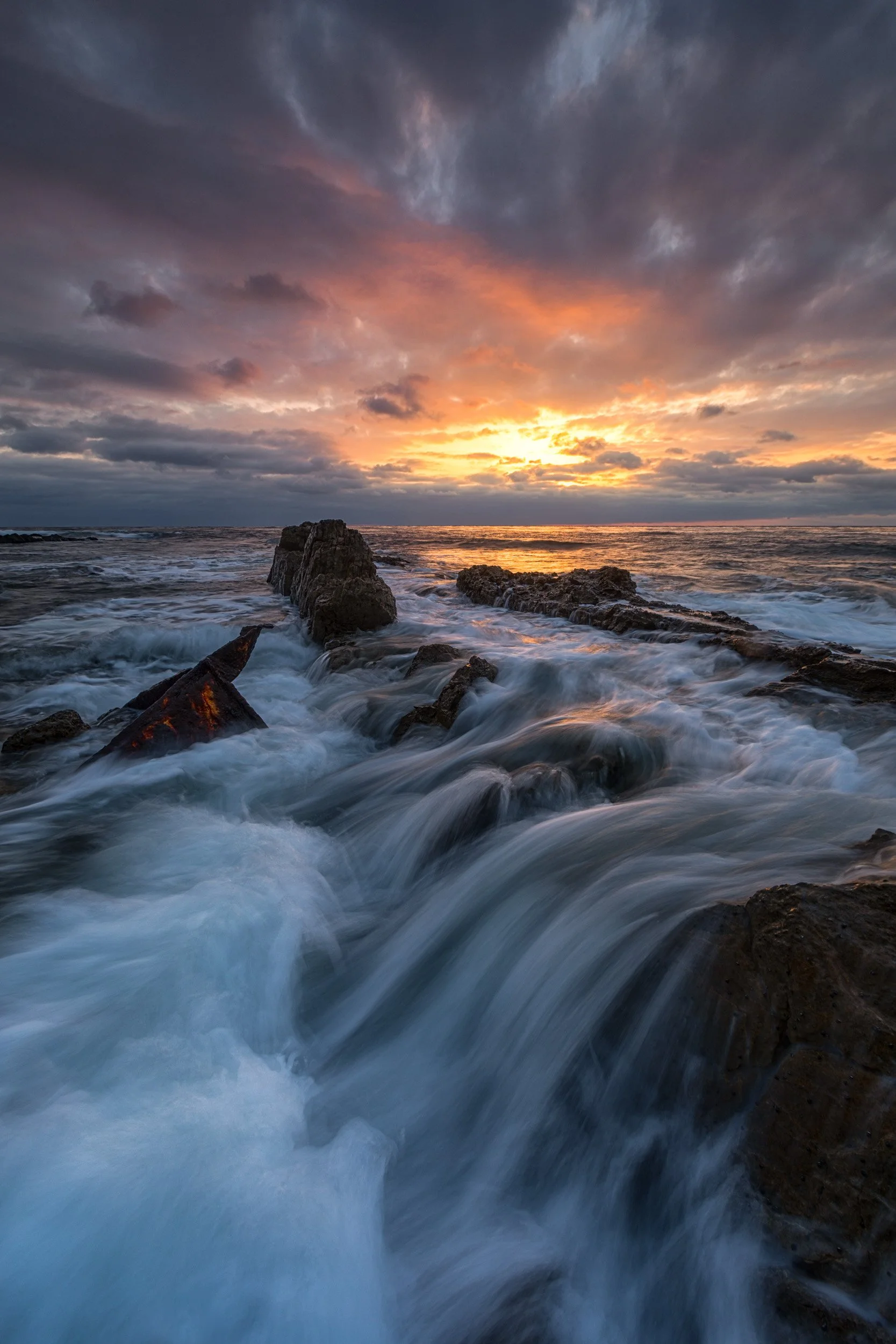 Vertical long exposure of rushing whitewater surging around the rusted S.S. Dominator shipwreck on the rocky Palos Verdes coast under a dramatic cloud-filled sunset by Chris Ewen Crosby.