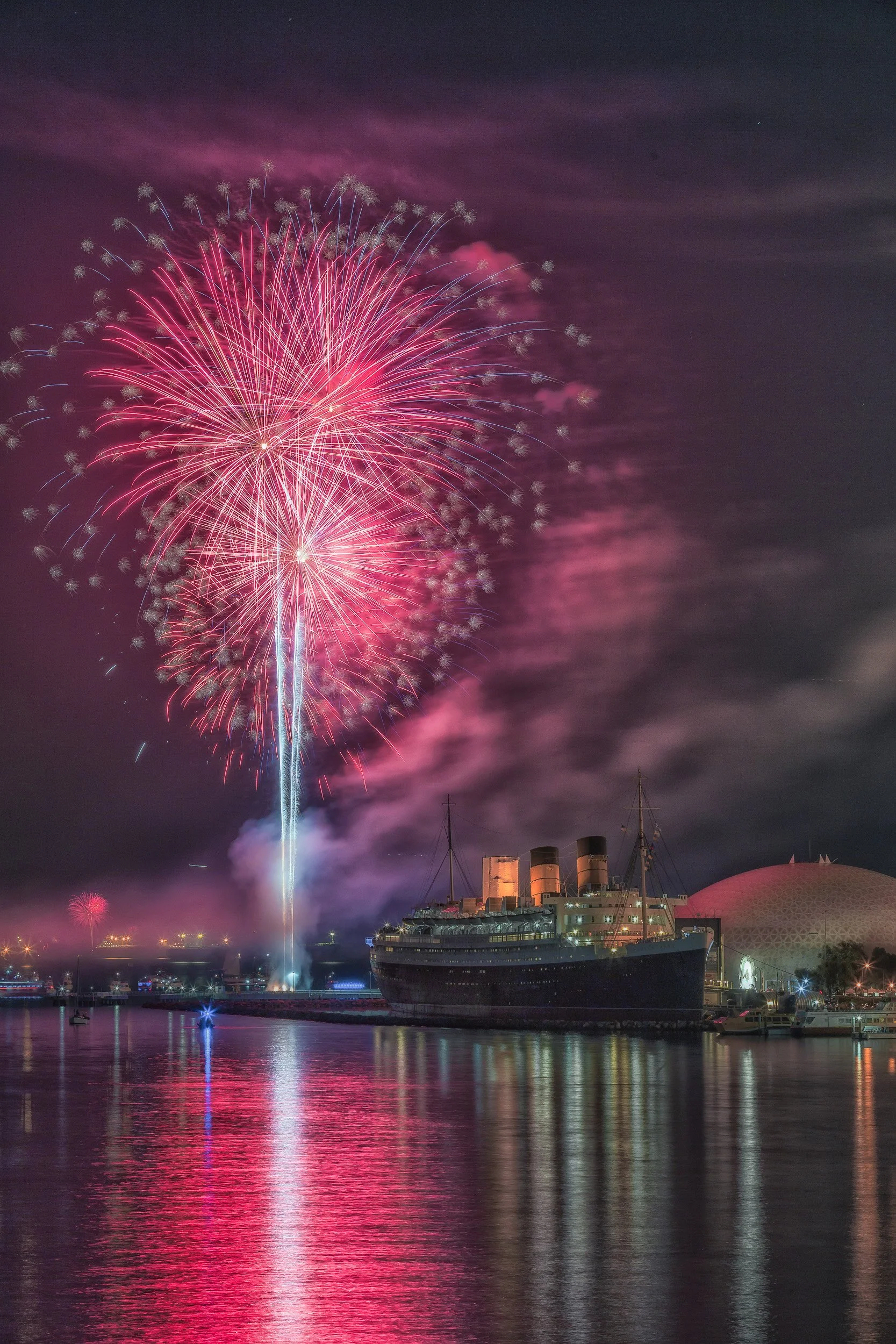 Vibrant pink fireworks exploding over the RMS Queen Mary and Long Beach harbor with water reflections by Chris Ewen Crosby.