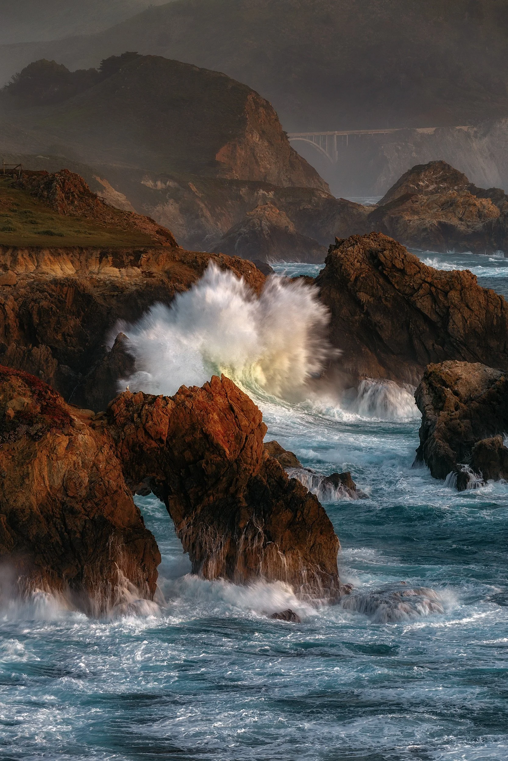 Vertical shot of a massive ocean wave exploding against rocky sea stacks along the Big Sur coastline with Bixby Creek Bridge in the distant background by Chris Ewen Crosby.