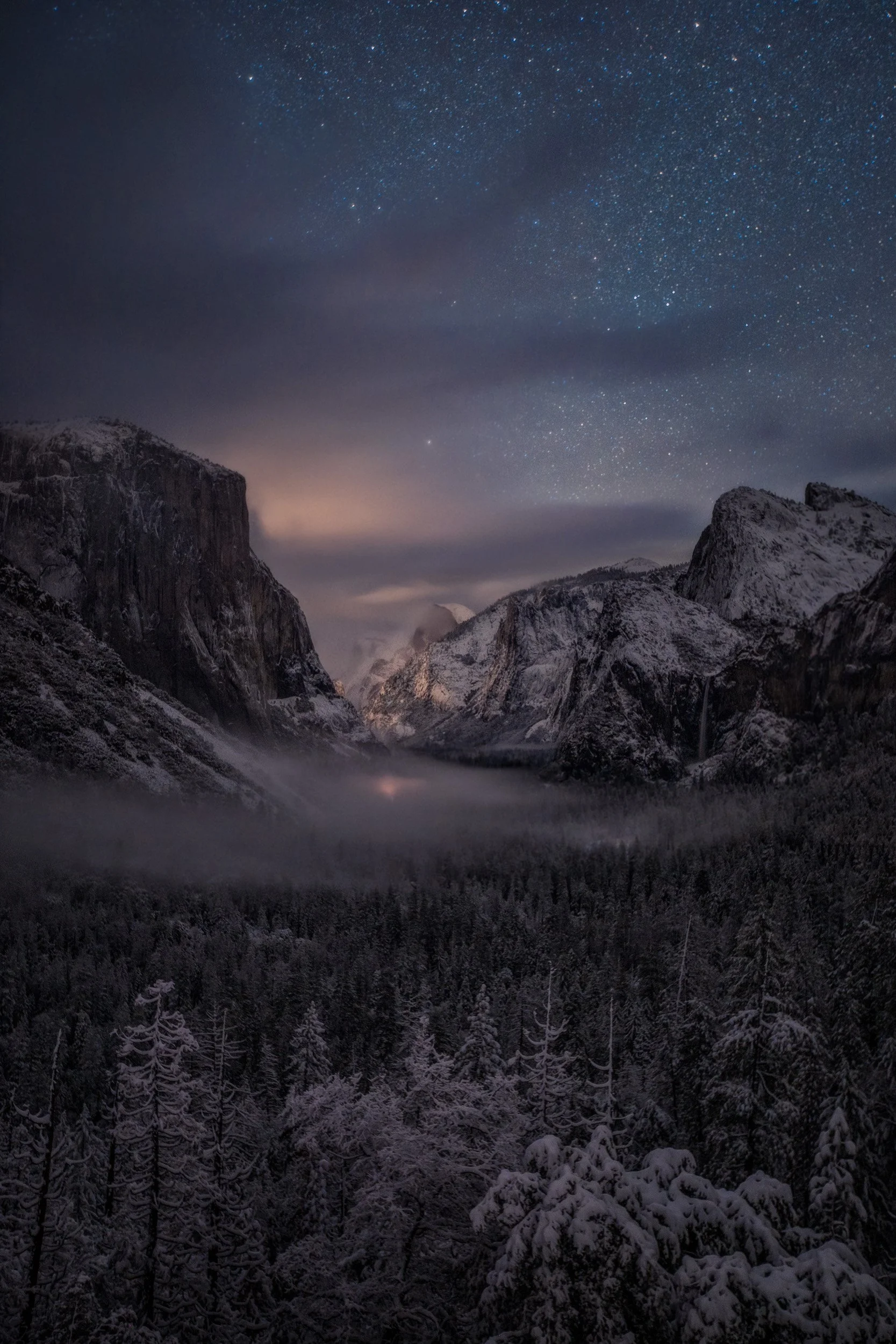 Vertical night landscape photograph of snow-covered pine trees and low valley fog framing El Capitan and Half Dome under a starry winter sky at Tunnel View in Yosemite National Park by Chris Ewen Crosby.