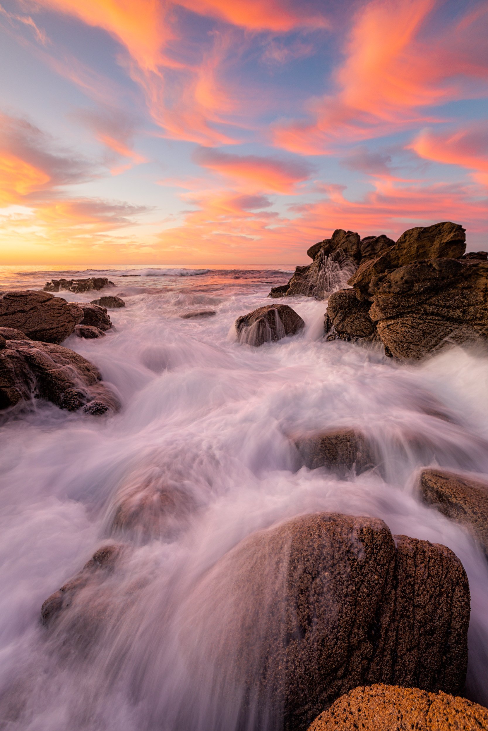 Vertical long exposure of surging ocean whitewater weaving through rocky shoreline outcrop beneath pink feathered sunset clouds in Laguna Beach by Chris Ewen Crosby.
