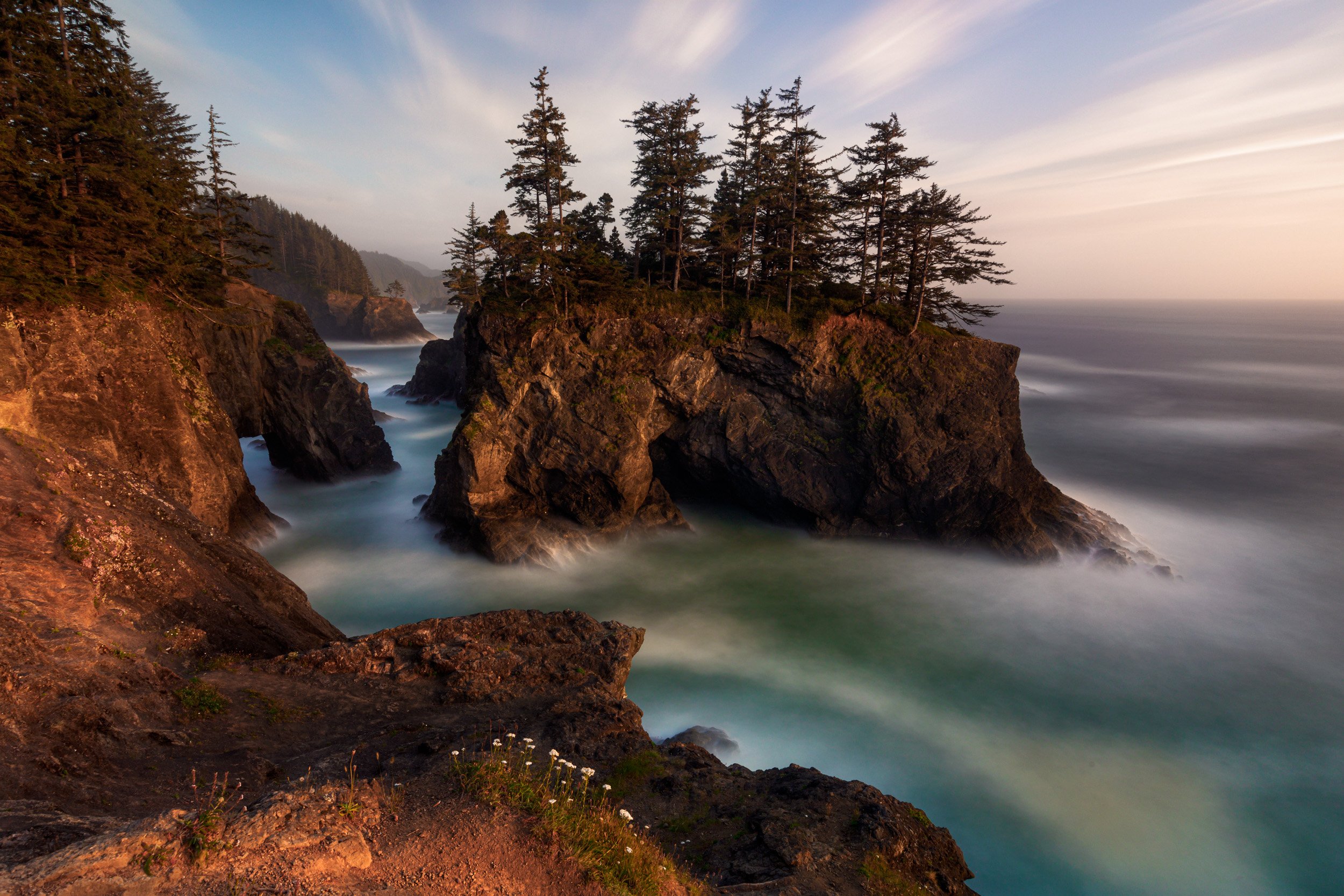 Horizontal long exposure photograph of tree-covered rock arches and natural bridges surrounded by mist-like ocean water along the Oregon Coast by Chris Ewen Crosby.