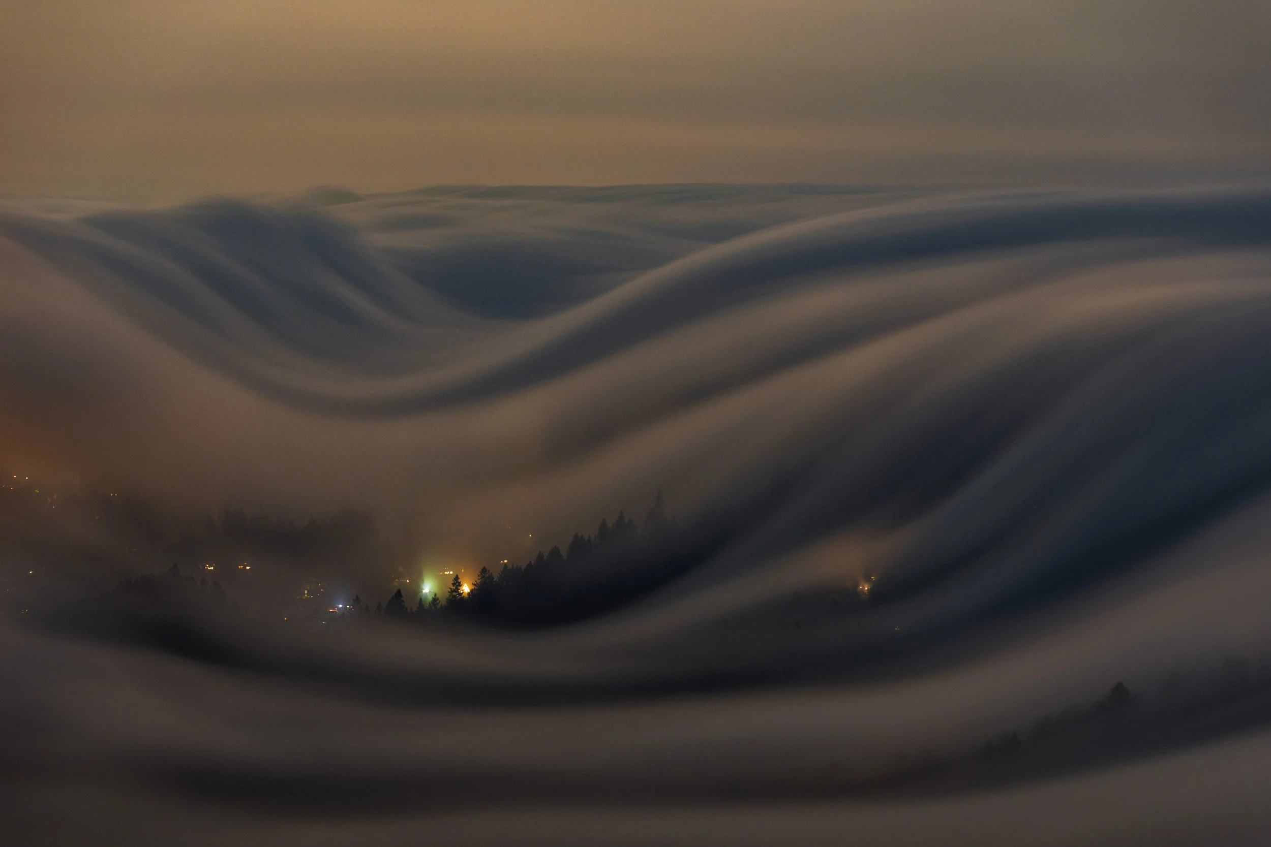Horizontal night long exposure landscape photograph of silky fog waves rolling over dark hills with glowing city lights peeking through the mist from Mount Tamalpais in Marin County by Chris Ewen Crosby.