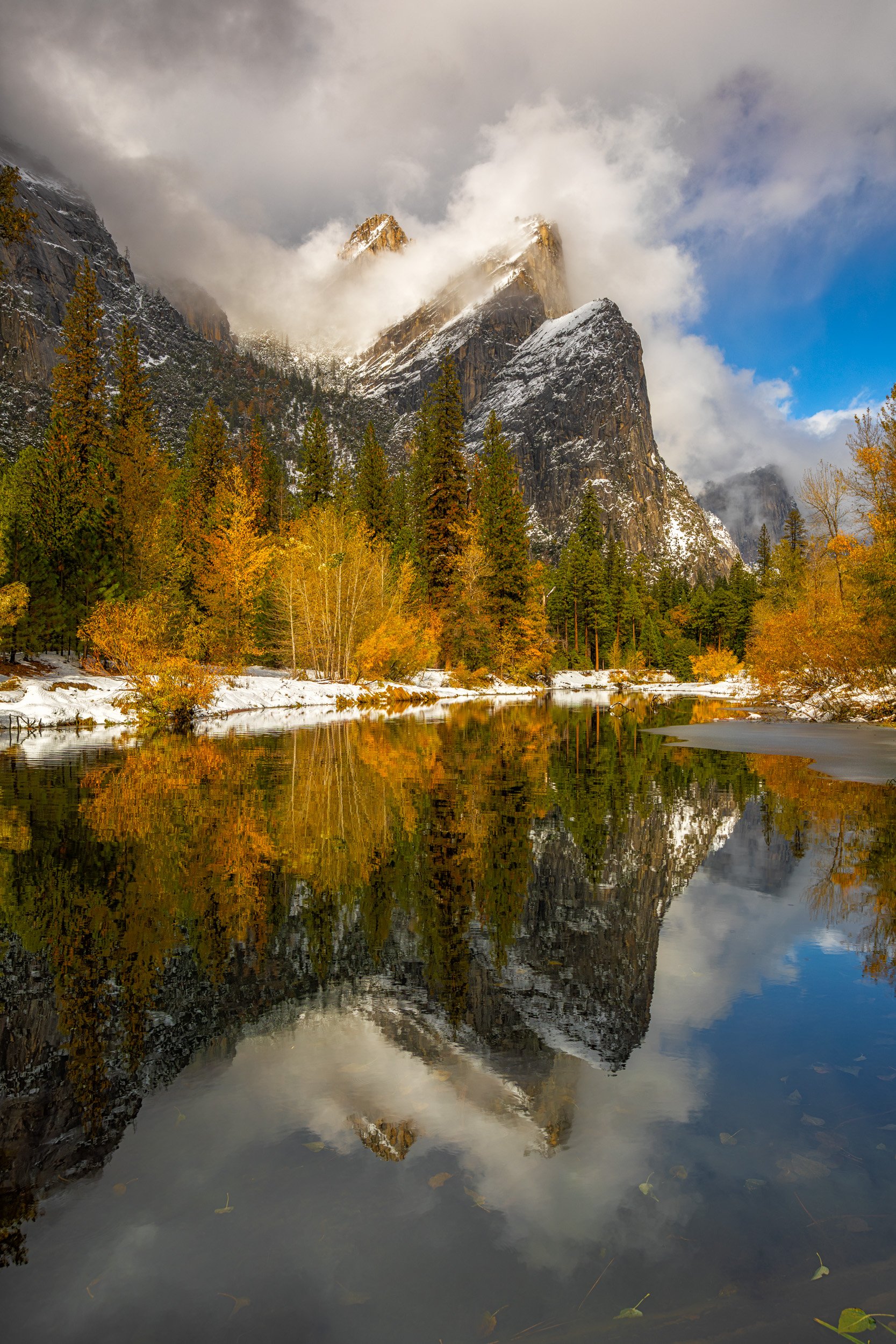 Vertical landscape photograph of snow-capped mountains and colorful autumn trees perfectly reflecting in a glassy river in Yosemite National Park.