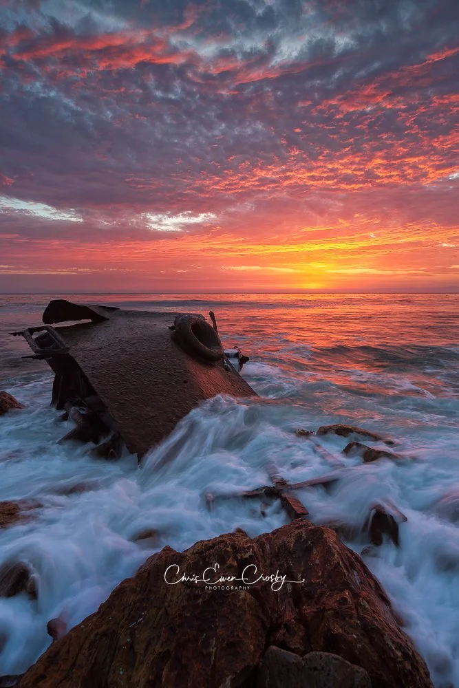 Long exposure sunset photography of a rusted shipwreck on a rocky shoreline in Palos Verdes, California, under a vibrant orange and yellow sky.