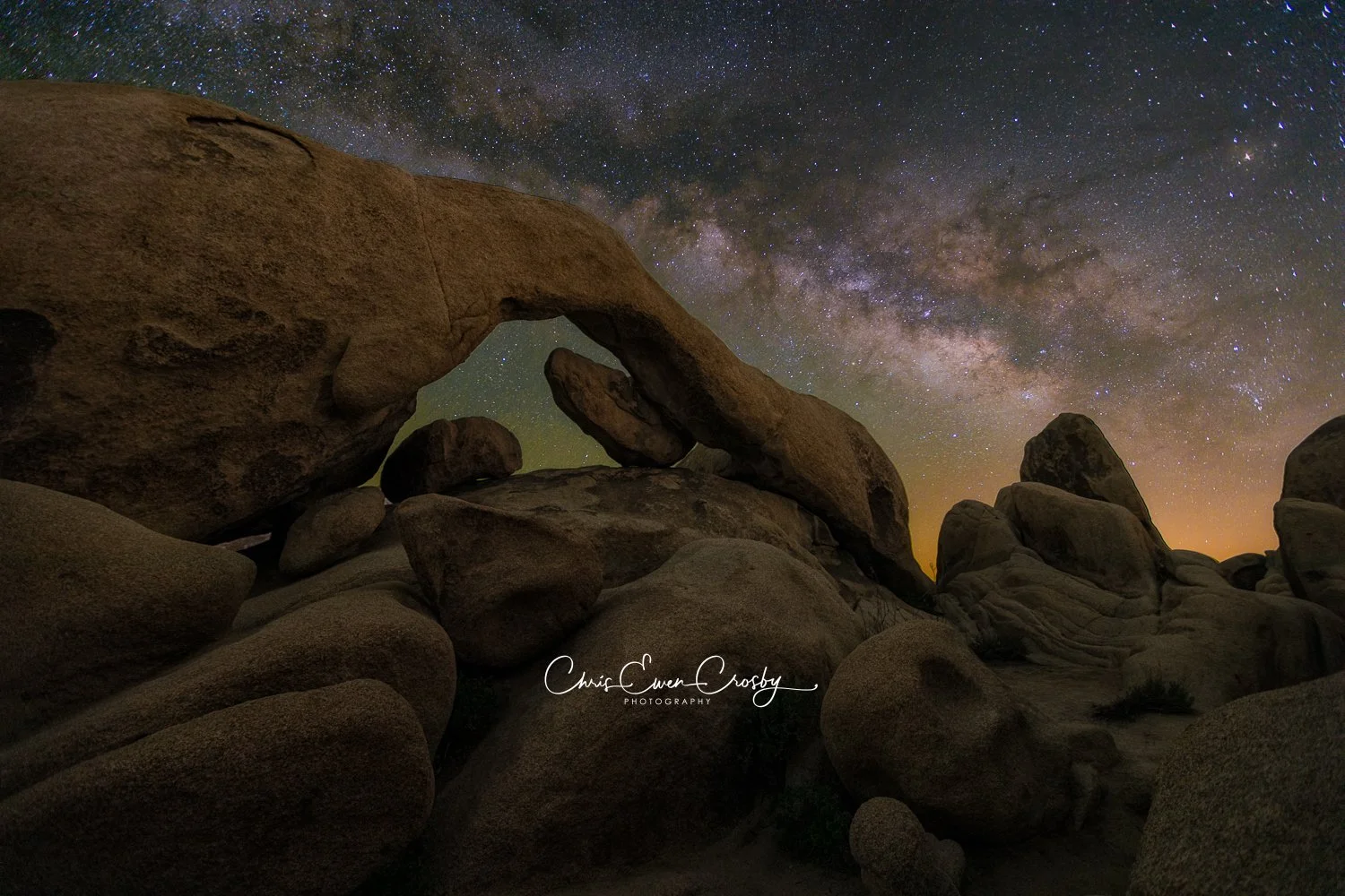 A horizontal landscape night photograph of a natural stone arch in Joshua Tree National Park with the bright Milky Way galaxy core visible in the starry sky above.