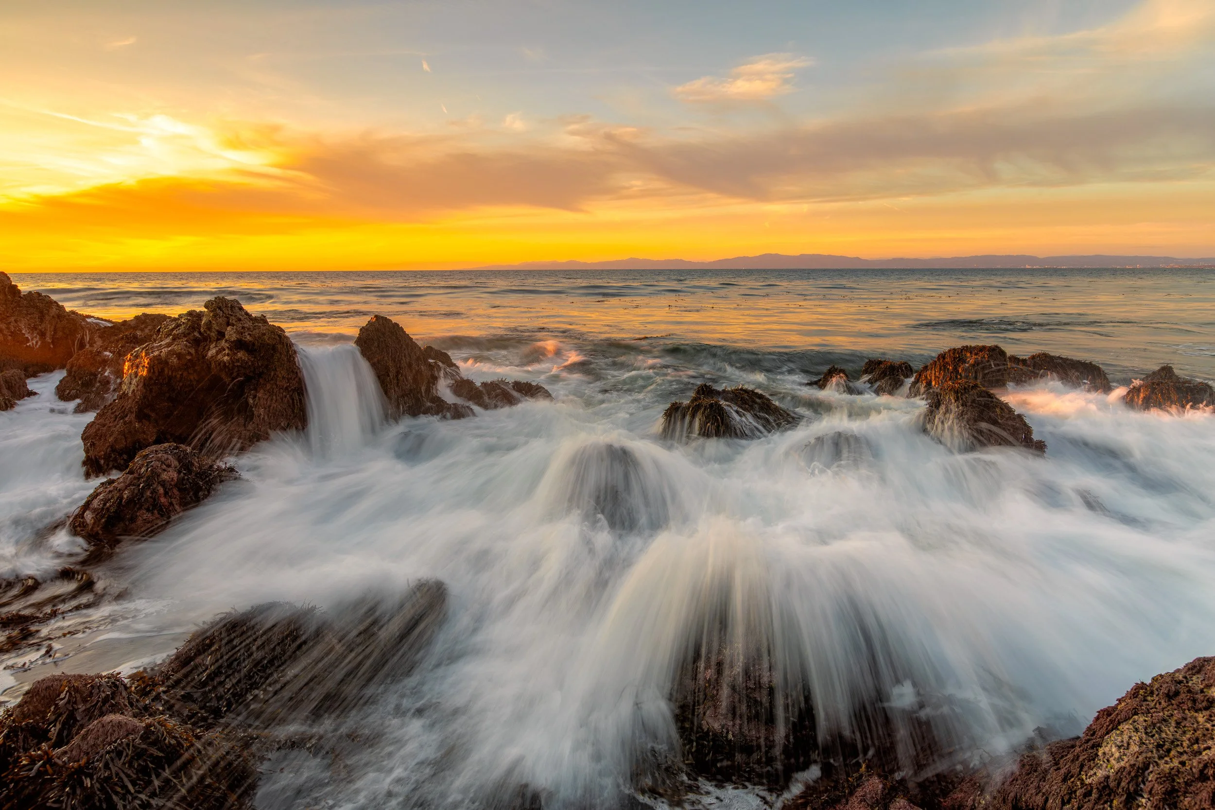 Horizontal long exposure photograph of ocean waves splashing over rocky outcroppings along the Palos Verdes coastline under a vibrant golden sunset sky by Chris Ewen Crosby.
