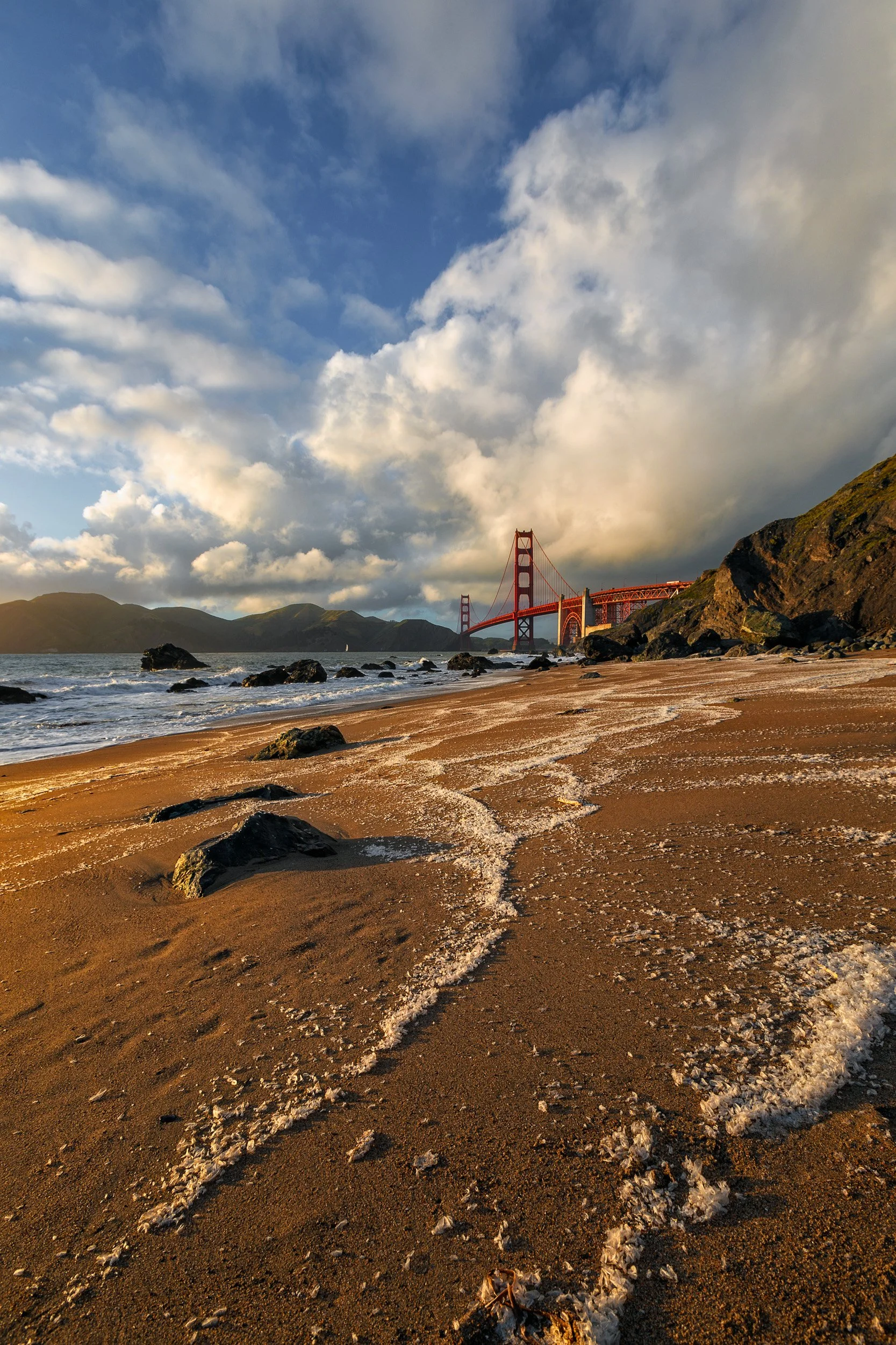 Vertical shot of sea foam patterns on the shoreline at Marshall's Beach looking toward the Golden Gate Bridge under dramatic clouds by Chris Ewen Crosby.