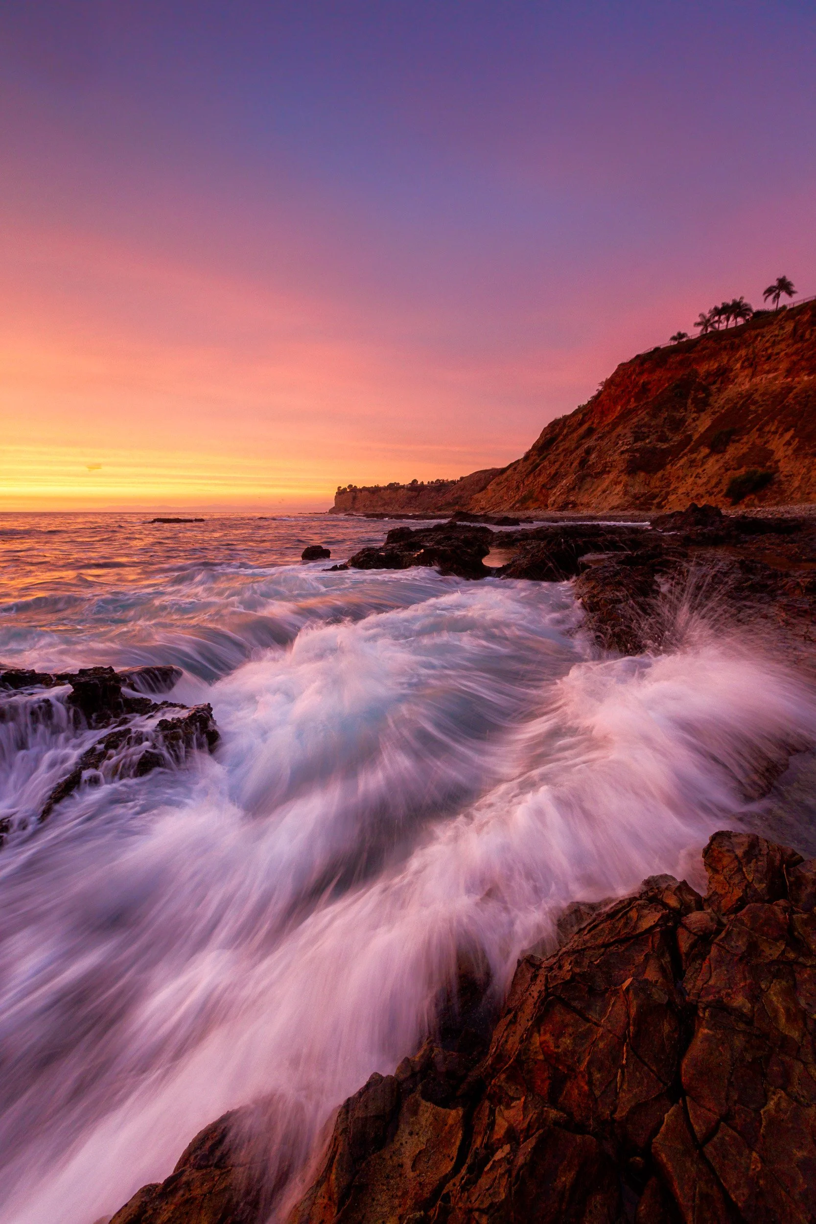 Vertical long exposure of churning waves sweeping over rocky coast beneath the steep Palos Verdes cliffs at sunset by Chris Ewen Crosby.
