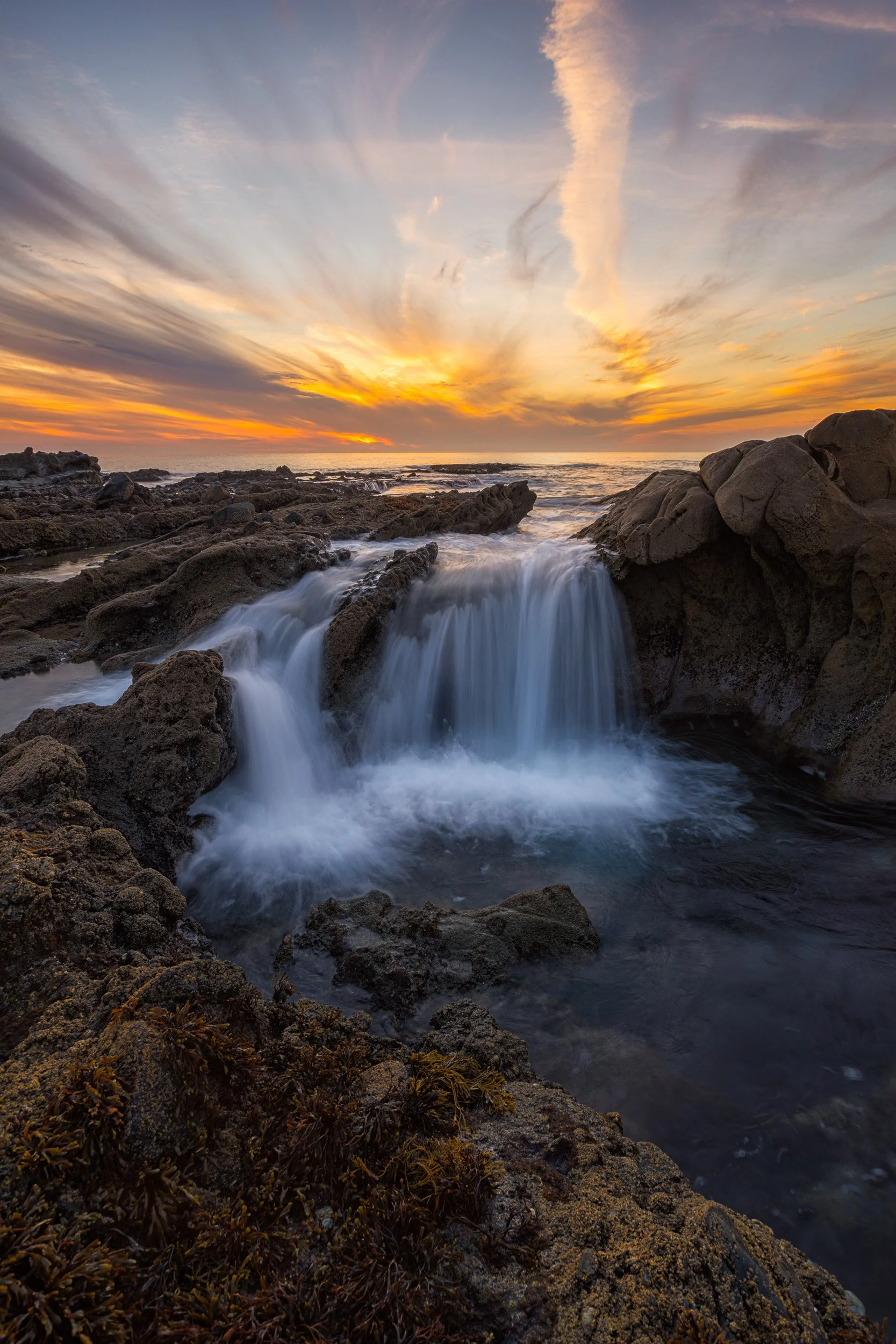 Vertical long exposure photograph of ocean water cascading like a waterfall into a rocky Laguna Beach tide pool under a golden sunset by Chris Ewen Crosby.