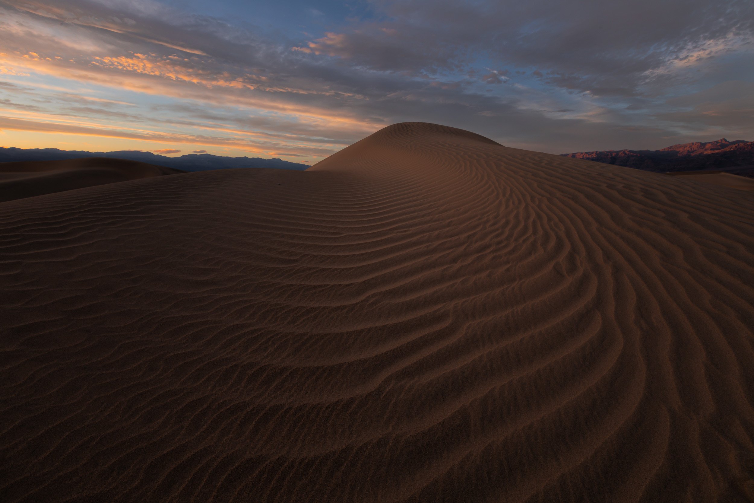 Horizontal landscape photograph of intricate wind-blown sand ripples sweeping across the Mesquite Flat Sand Dunes beneath a pastel sunset sky in Death Valley National Park by Chris Ewen Crosby.
