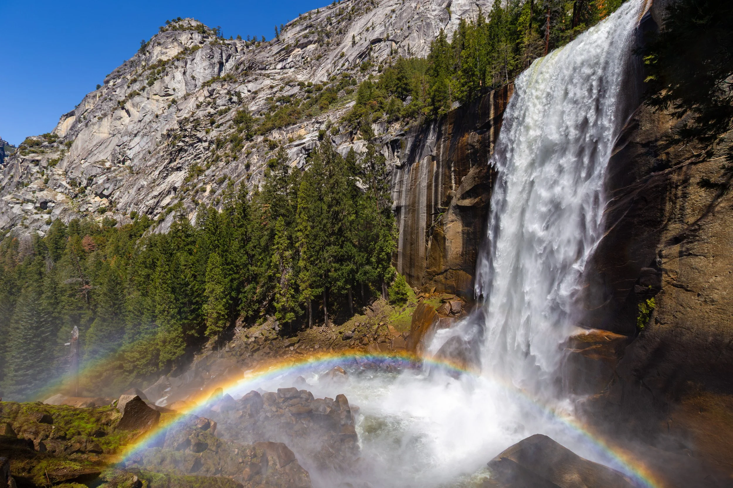 Wide-angle landscape photograph of Vernal Fall in Yosemite National Park during peak spring snowmelt, featuring a bright, full rainbow arching through the misty spray below sheer granite cliffs.