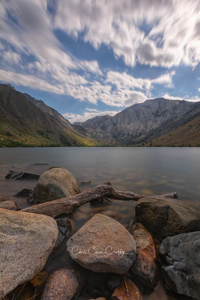 Long exposure landscape of Convict Lake with calm water, rugged mountain peaks, and soft white clouds at afternoon.