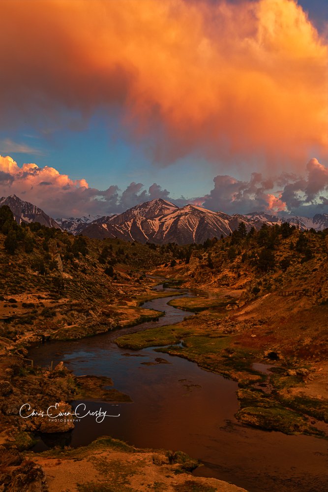 Vertical photo of a bright pink and purple sunset over a steaming turquoise creek in a rugged canyon at Hot Creek Geologic Site.