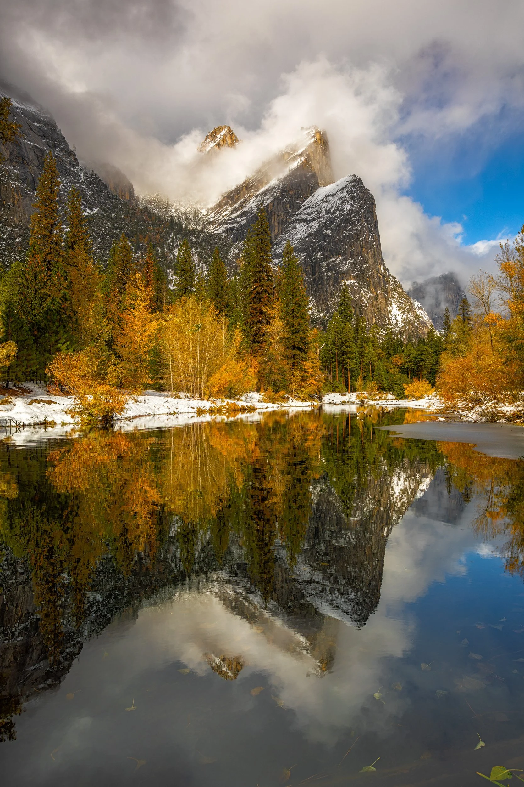 Vertical landscape photograph of golden autumn trees and snow-dusted Three Brothers granite peaks perfectly reflected in the Merced River as storm clouds clear in Yosemite National Park by Chris Ewen Crosby.