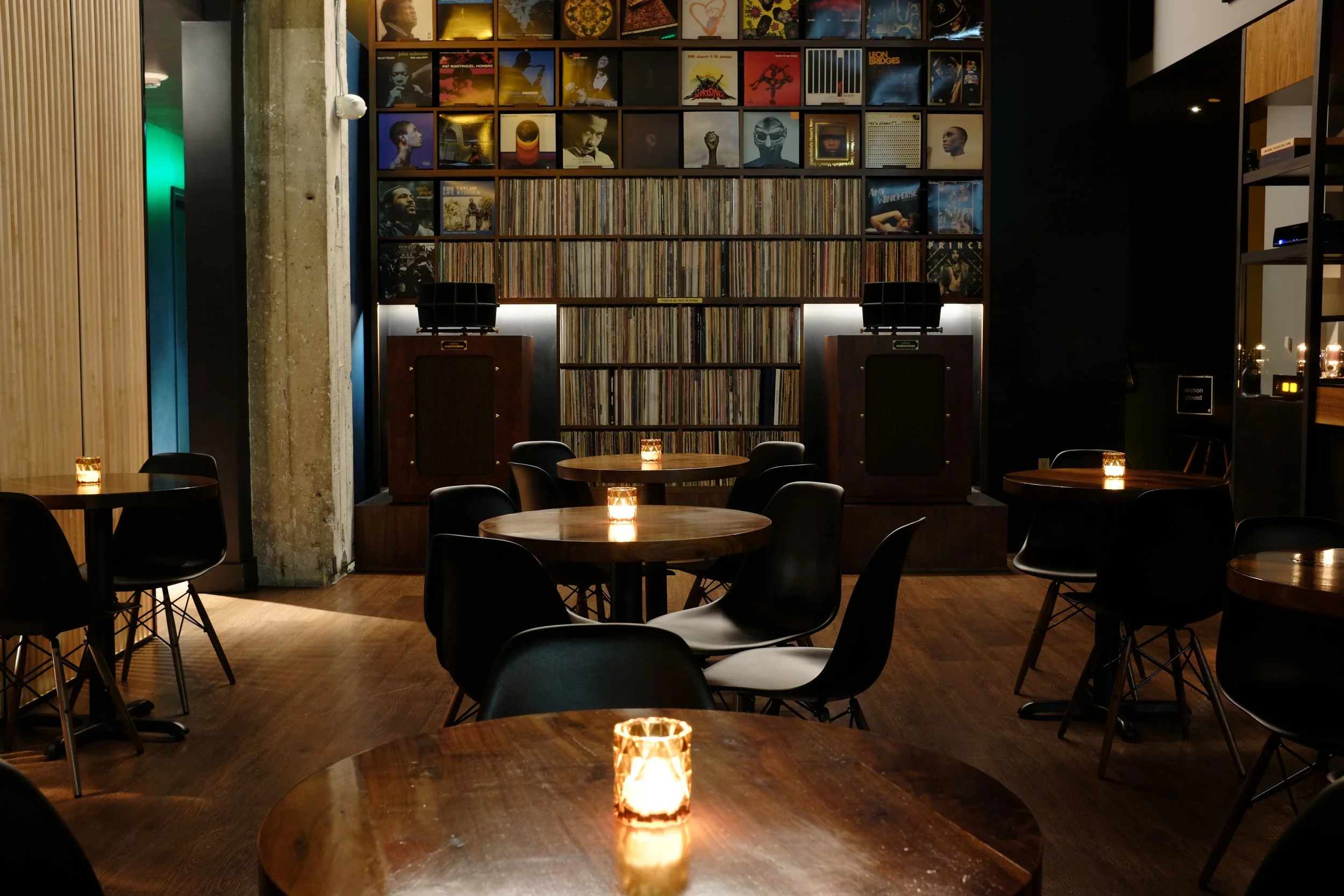 alt="Bar Shiru interior with small tables, with a large wall of vinyl records and high fidelity speakers in the background.""