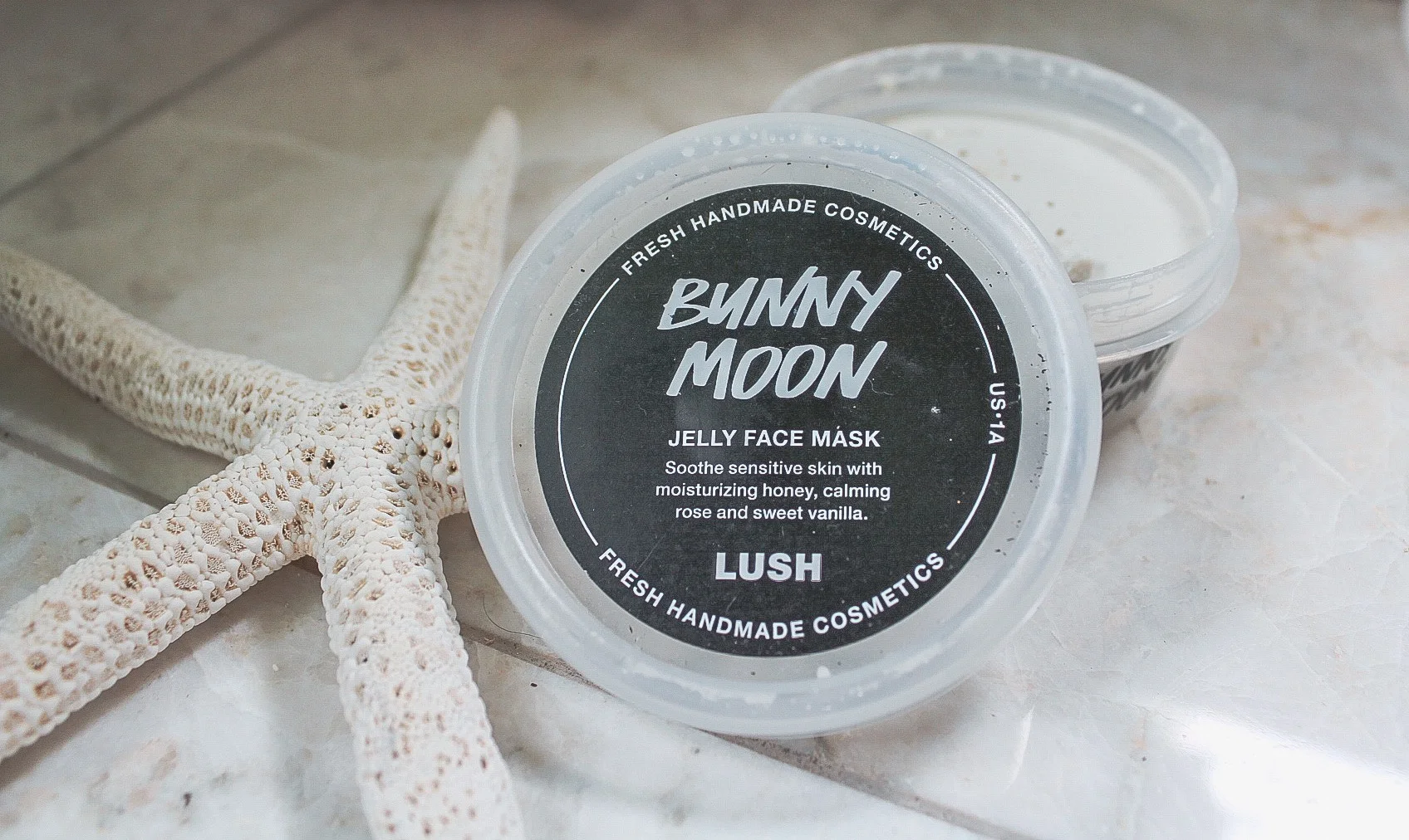 Photo description: A pot of Lush’s cream colored Bunny Moon jelly face mask is sitting on a marble counter open with the top leaning on it. There is a white starfish next to it.
